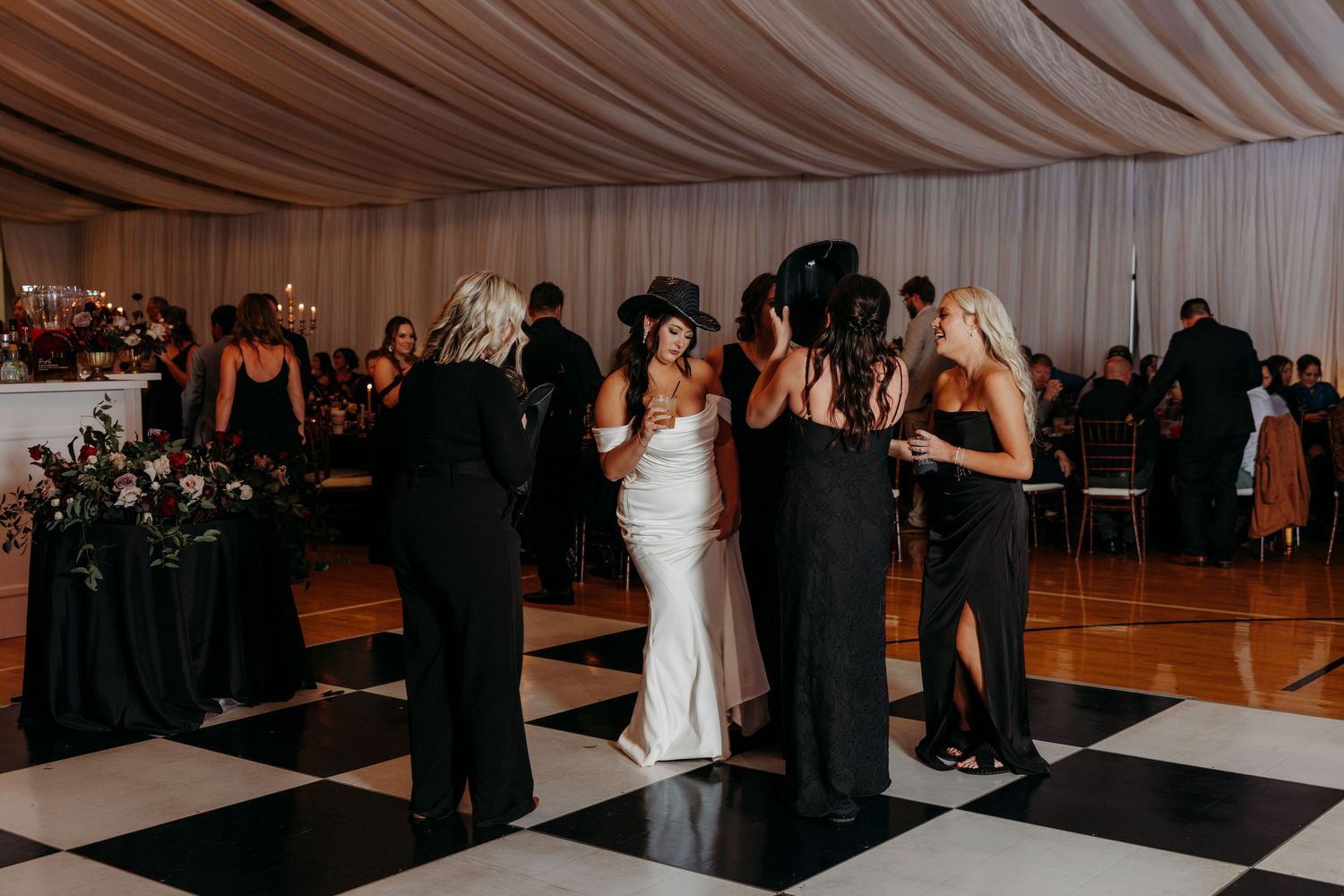Bride in white gown surrounded by bridesmaids in black dresses dancing on checkered black and white floor at wedding reception.