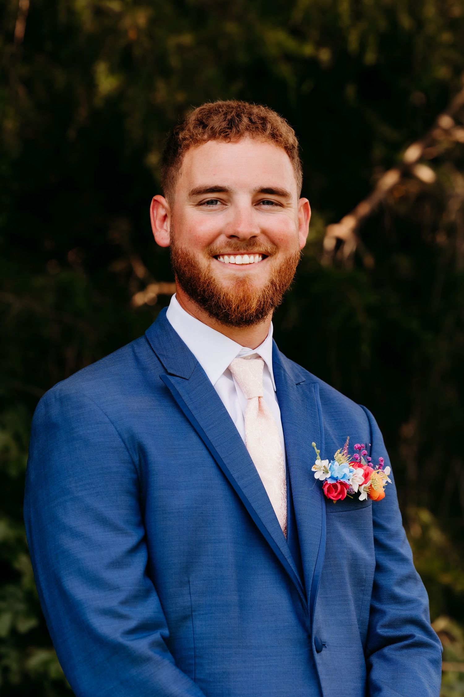 Smiling groom in blue suit with colorful boutonniere posing outdoors.