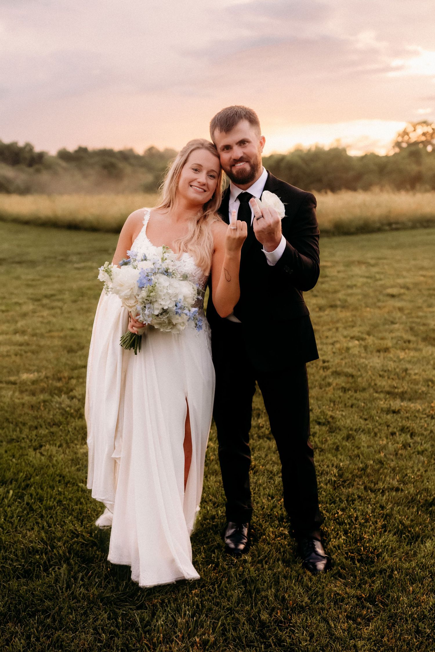 Bride and groom showing wedding rings at sunset, she holds blue hydrangea bouquet in white gown.