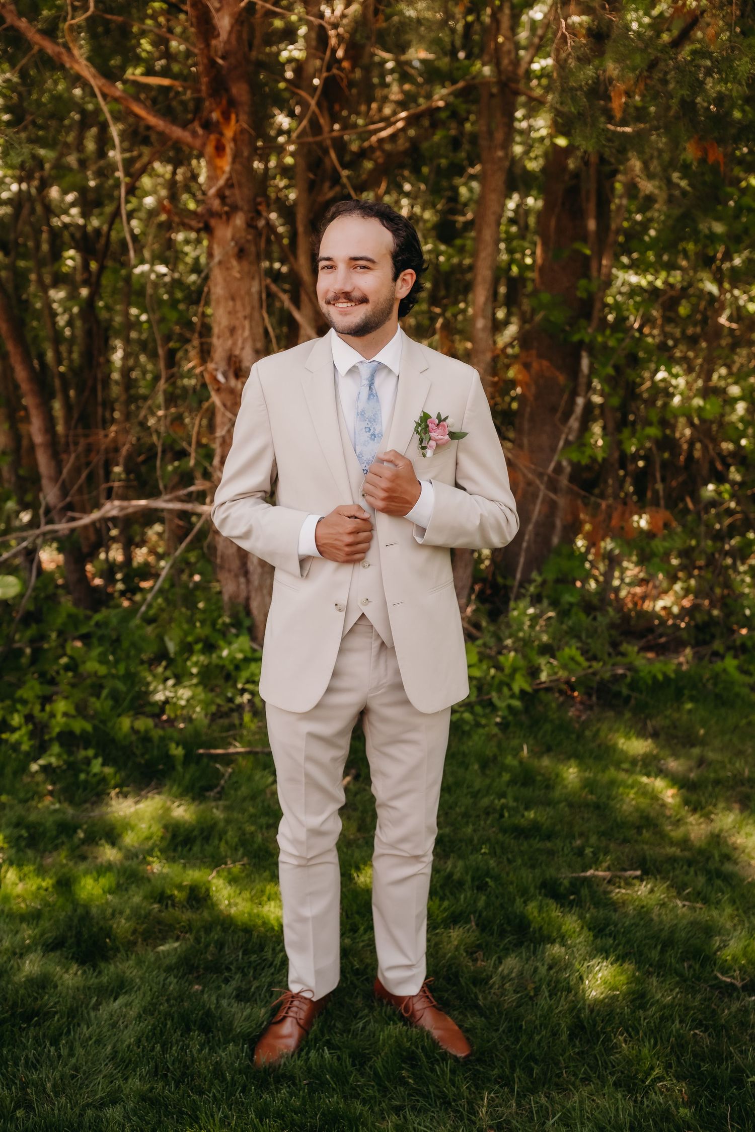 Smiling groom in beige three-piece suit with blue tie and floral boutonniere standing on green grass in a wooded area.