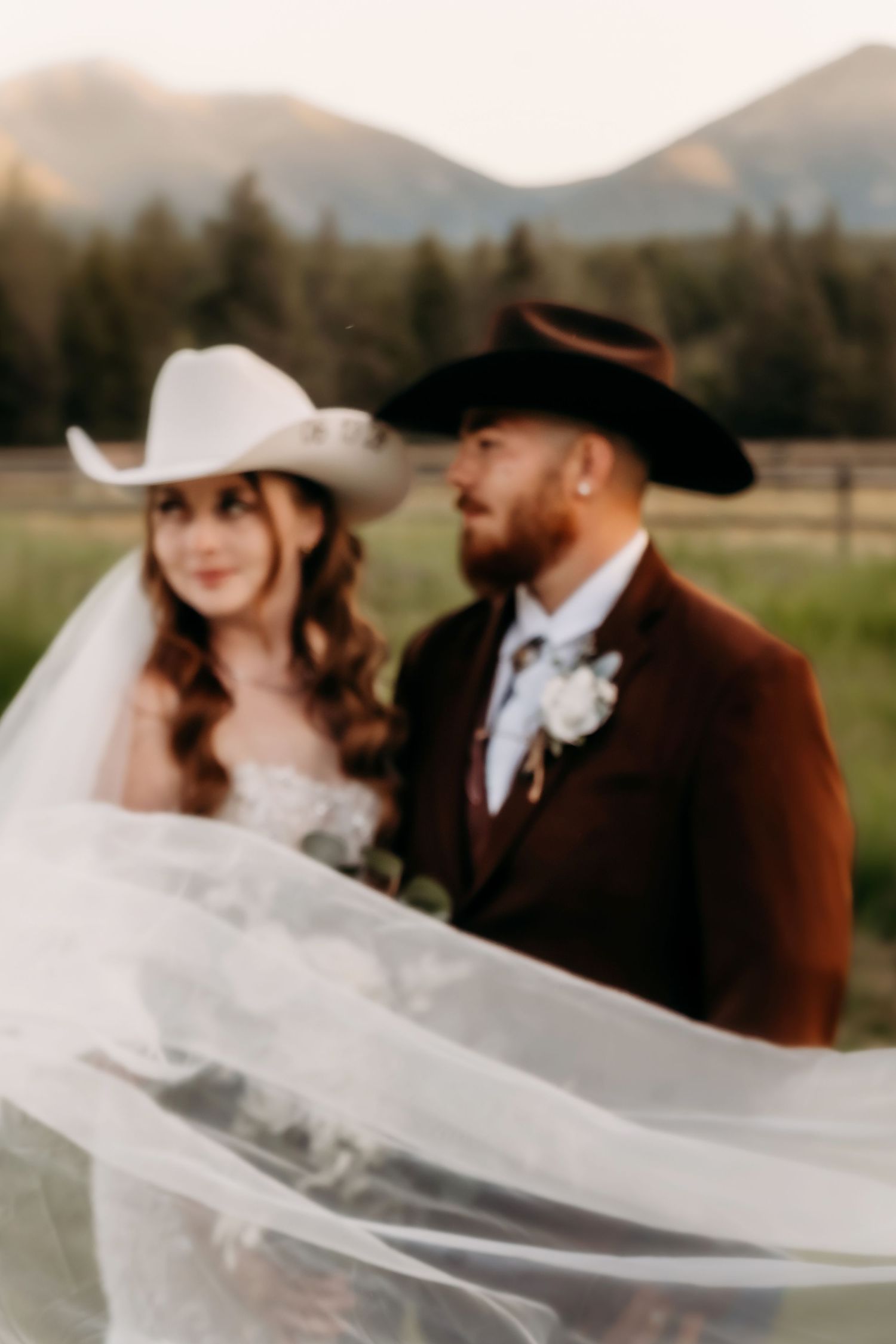 A bride and groom in cowboy hats pose outdoors with mountains and trees in the background on their wedding day.