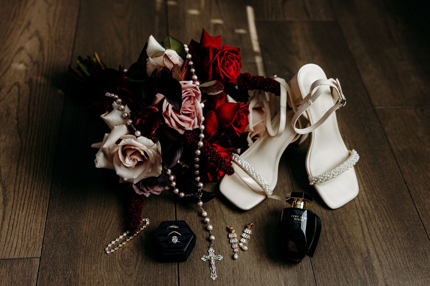 Bridal flat lay featuring white heeled sandals, red and pink roses bouquet, pearl rosary, cross pendant, and perfume bottles.