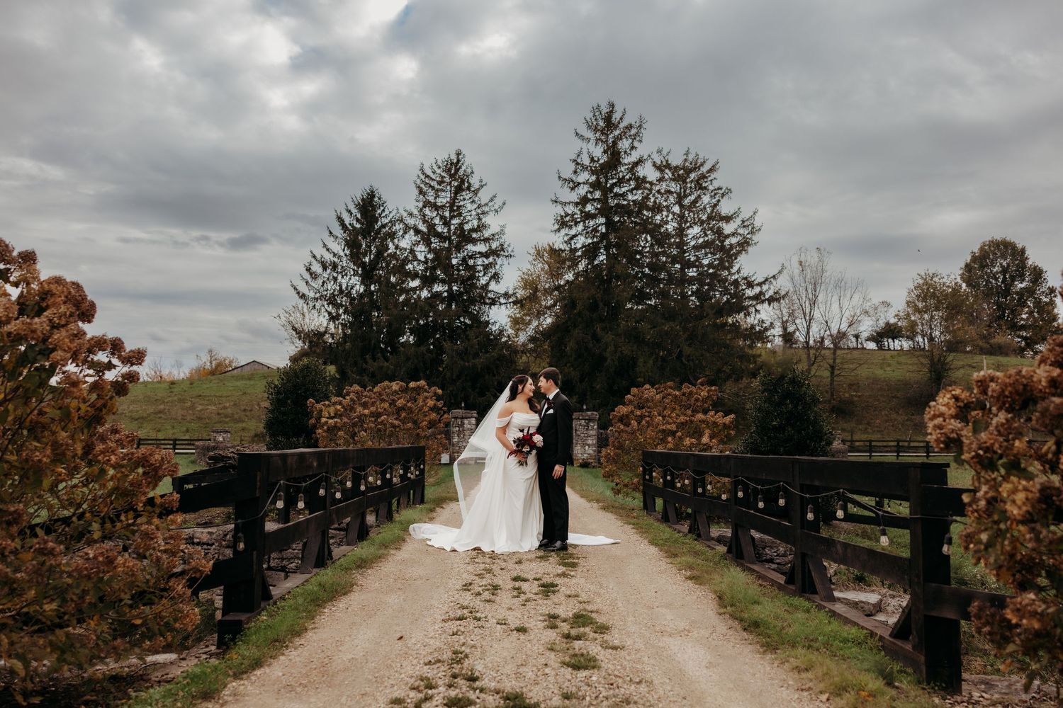Bride and groom share romantic moment on rustic fence-lined path surrounded by autumn trees under cloudy sky.