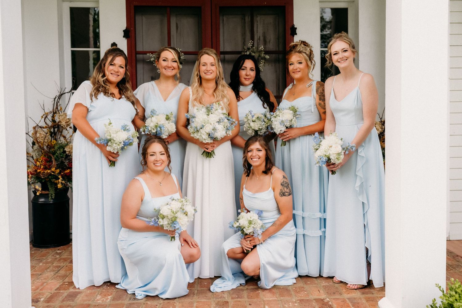 Seven bridesmaids in light blue dresses holding white floral bouquets pose on a white porch for a wedding photo.