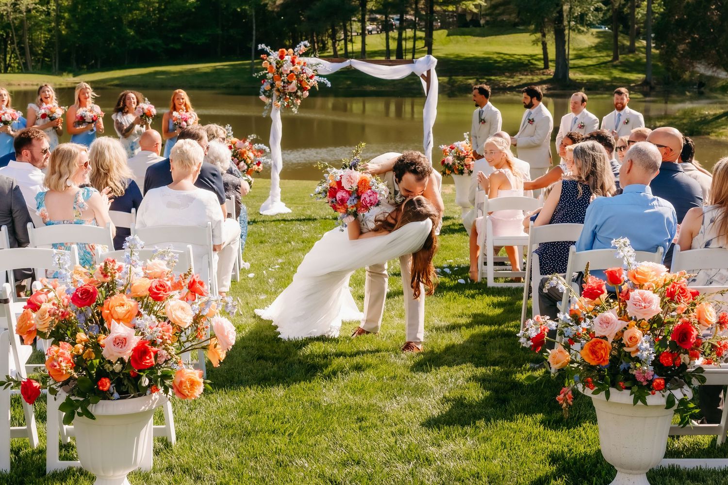 Couple shares first kiss at outdoor wedding ceremony with colorful floral arrangements and guests seated on white chairs.
