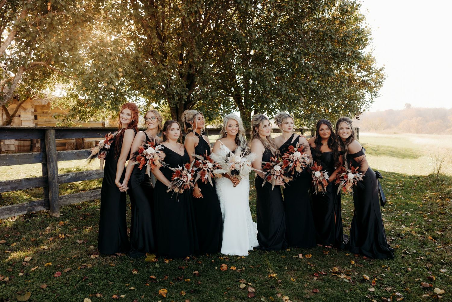 Wedding party in black dresses and white gown posing with autumn floral bouquets outside.