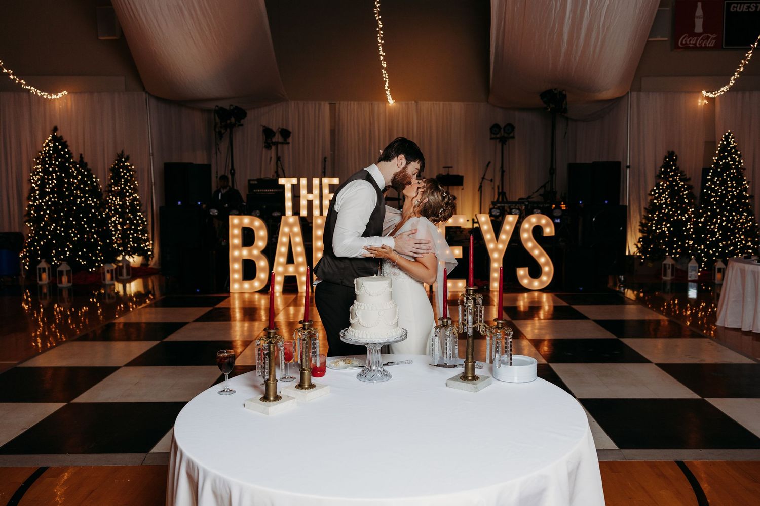 Newlyweds share intimate moment in front of lit marquee letters and Christmas trees.