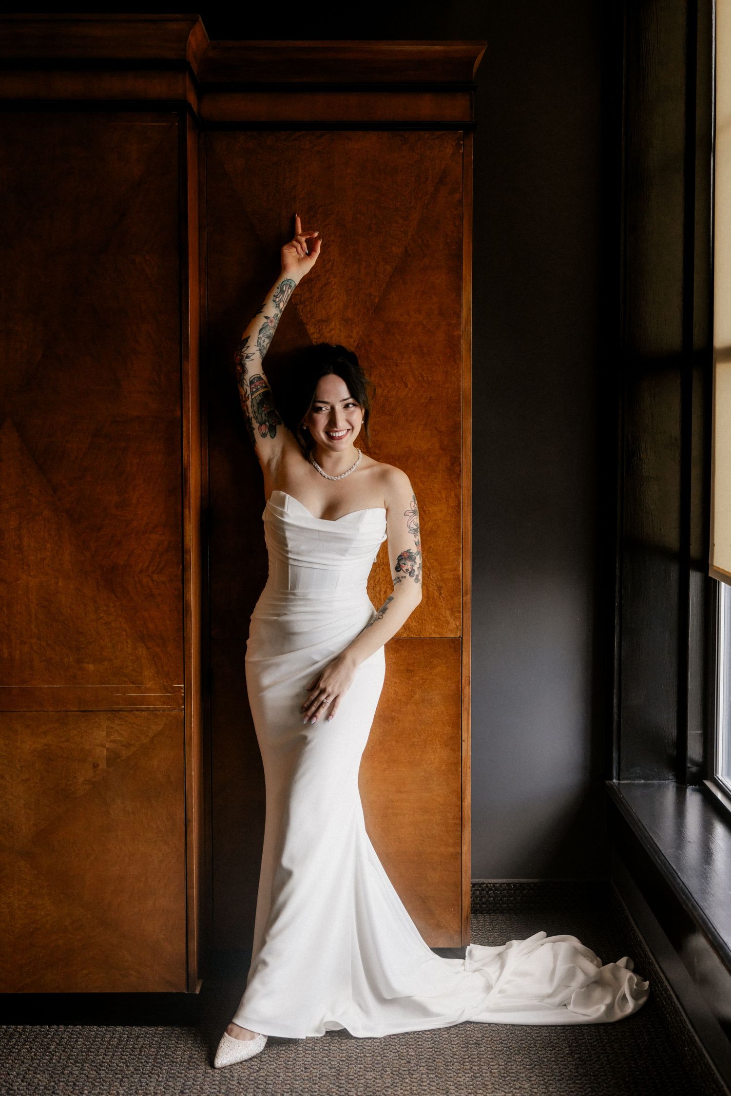 Bride in flowing white gown poses against warm wooden wall in natural light.