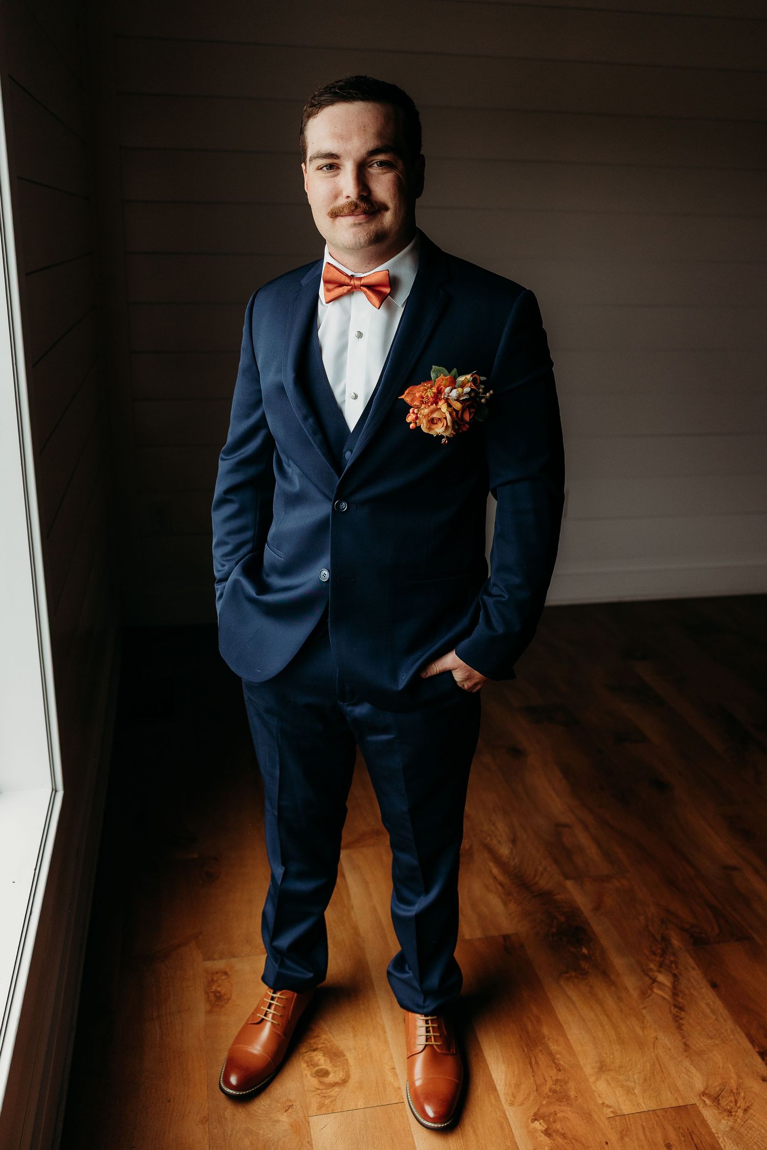 Groom in navy blue suit with orange bow tie and floral boutonniere poses indoors on hardwood floor.