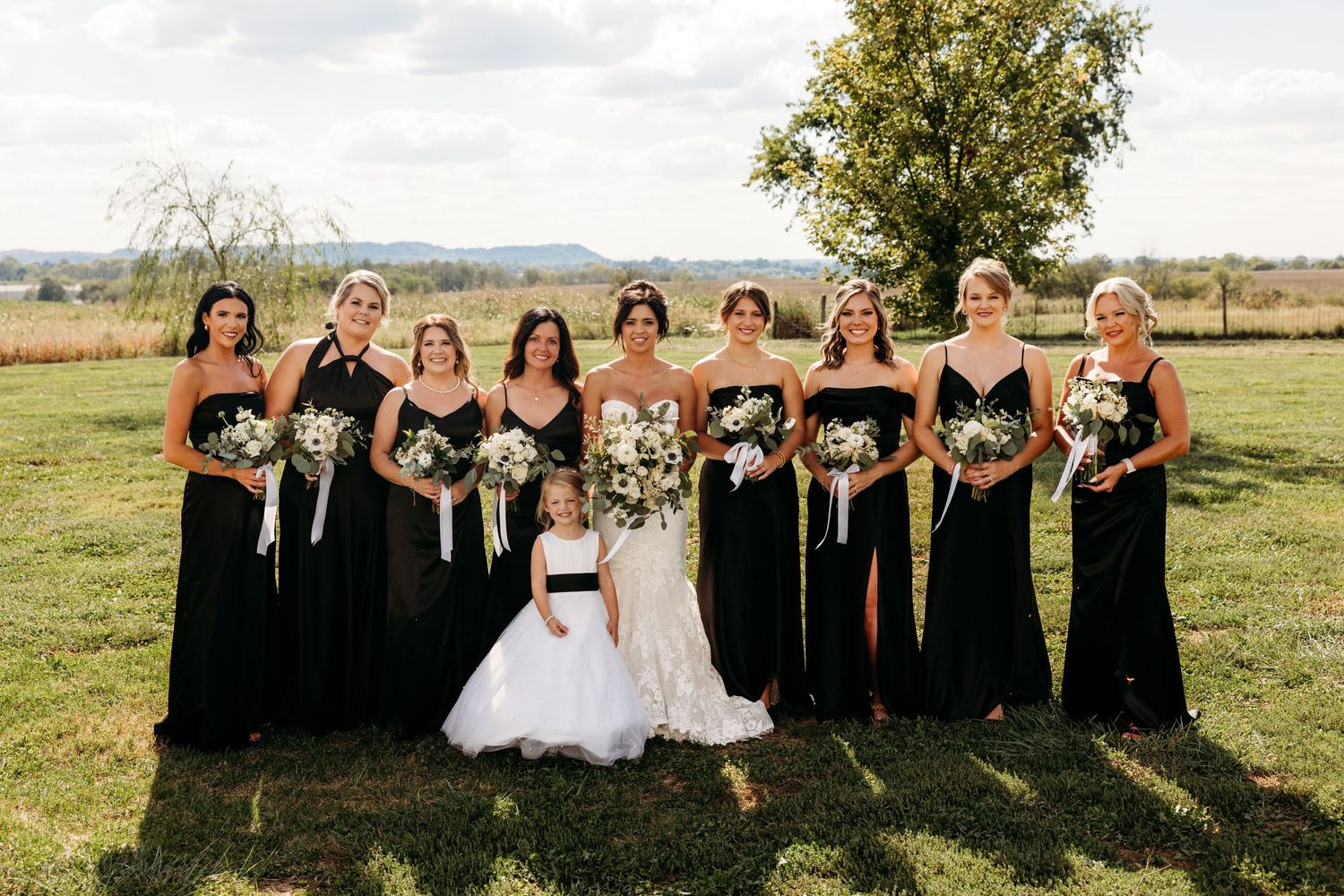 Bride in white gown with bridesmaids in black dresses and flower girl holding bouquets outdoors.