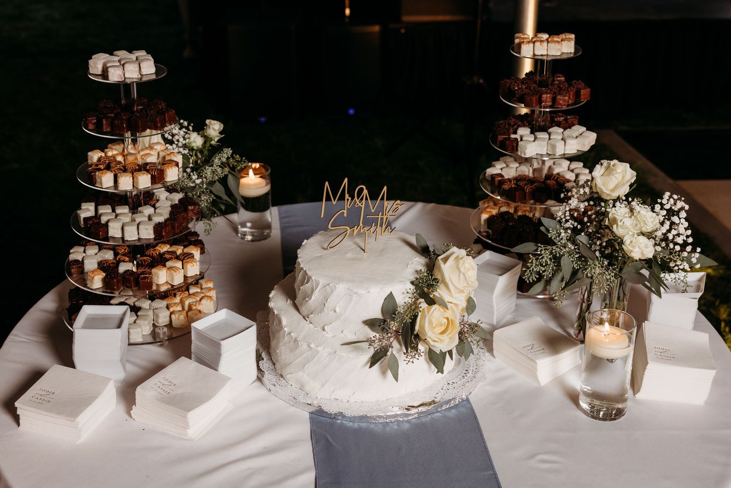 Elegant wedding dessert table featuring a white cake with florals, tiered treat towers, and candles.