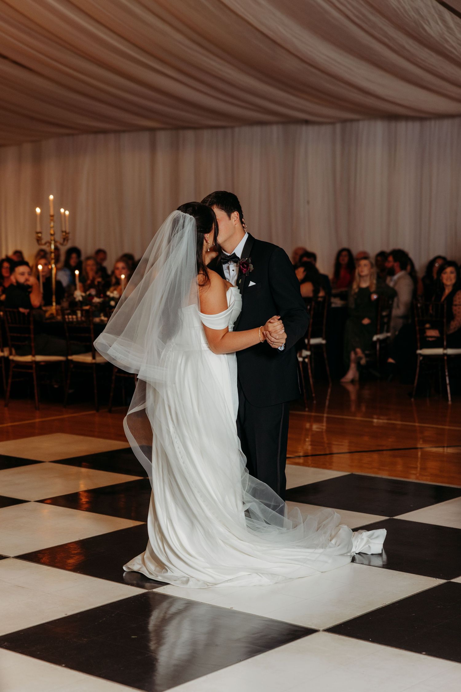 Bride and groom sharing first dance kiss on black and white checkered floor while wedding guests watch in elegant tented venue.