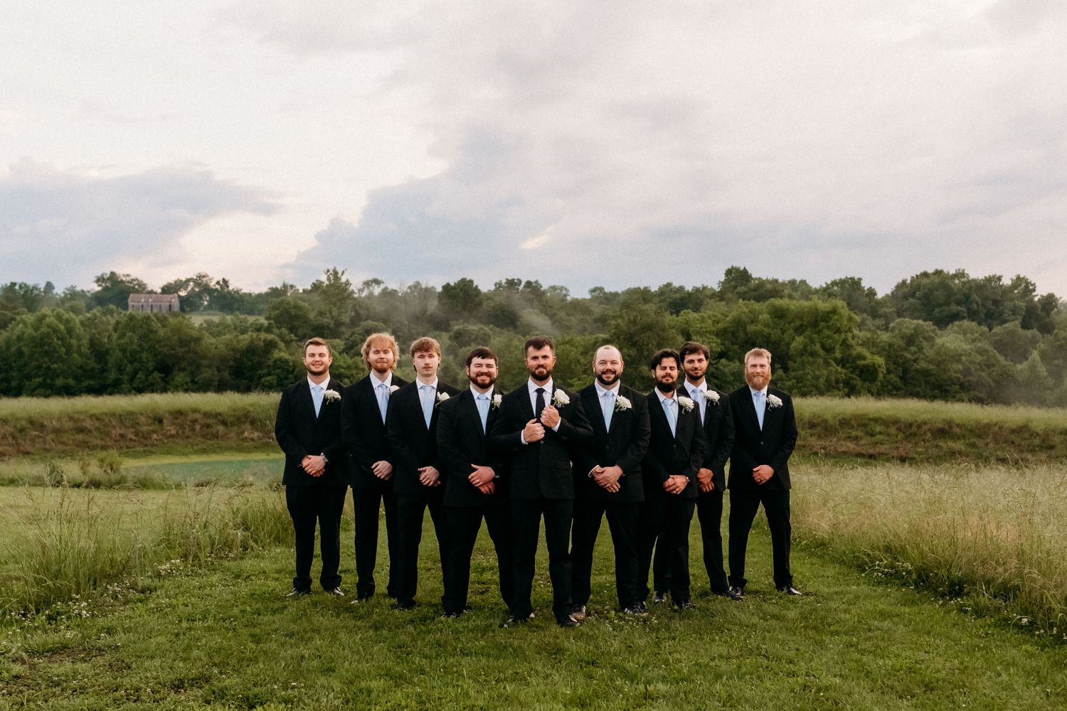 Groomsmen in black suits with white boutonnieres pose outdoors on green grass with trees and cloudy sky backdrop.