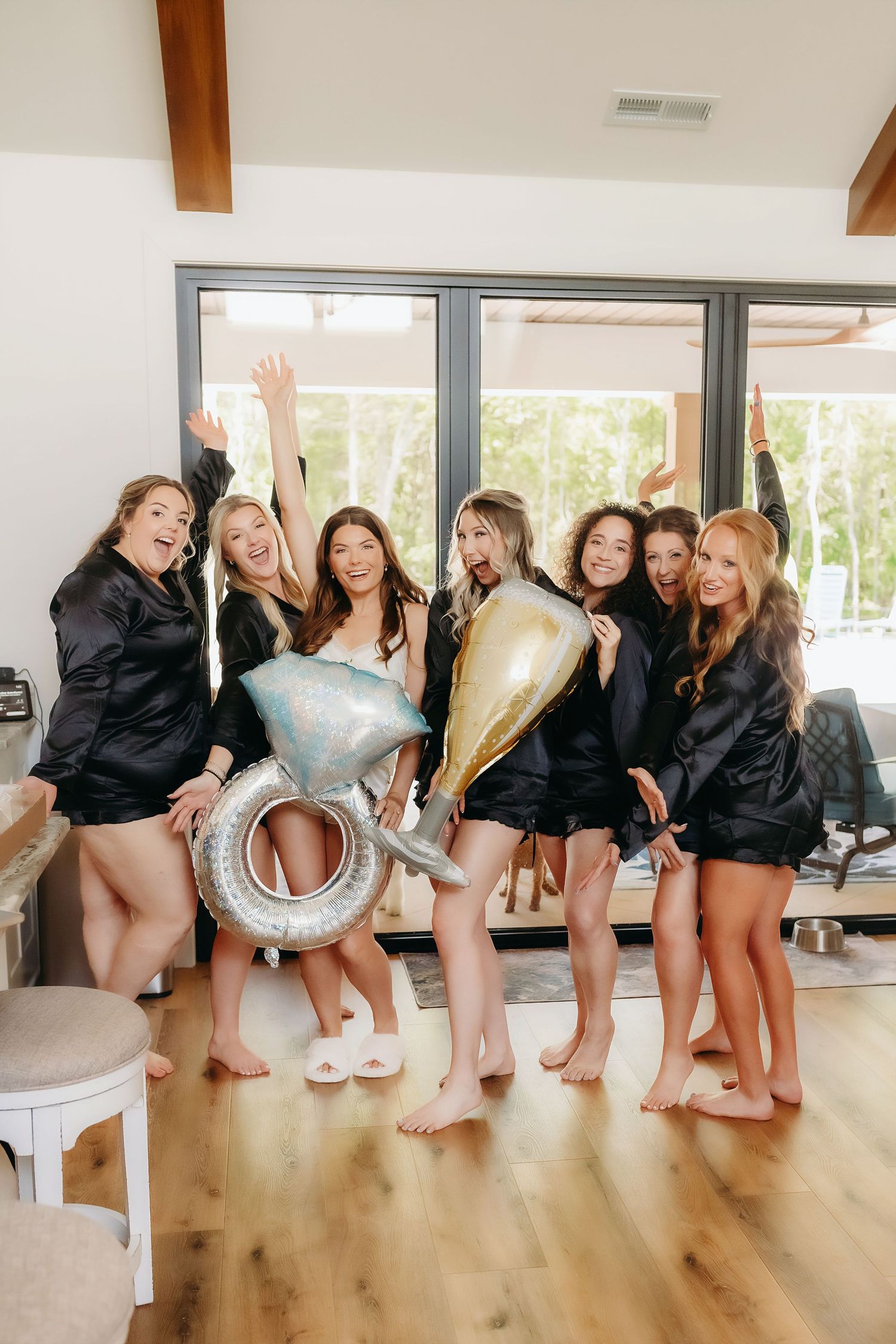 Bridesmaids in black satin robes celebrating with ring and champagne balloons during bridal party getting ready.