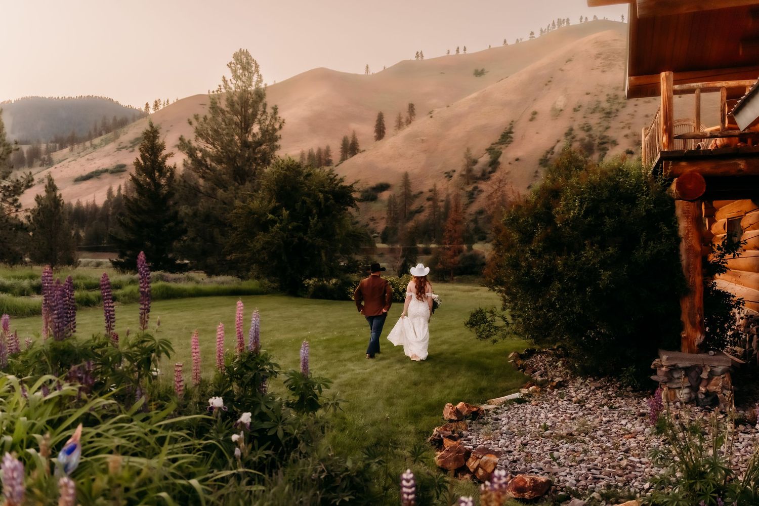 Couple walks hand-in-hand through lush garden with purple lupines, rustic cabin, and golden hillside at sunset.