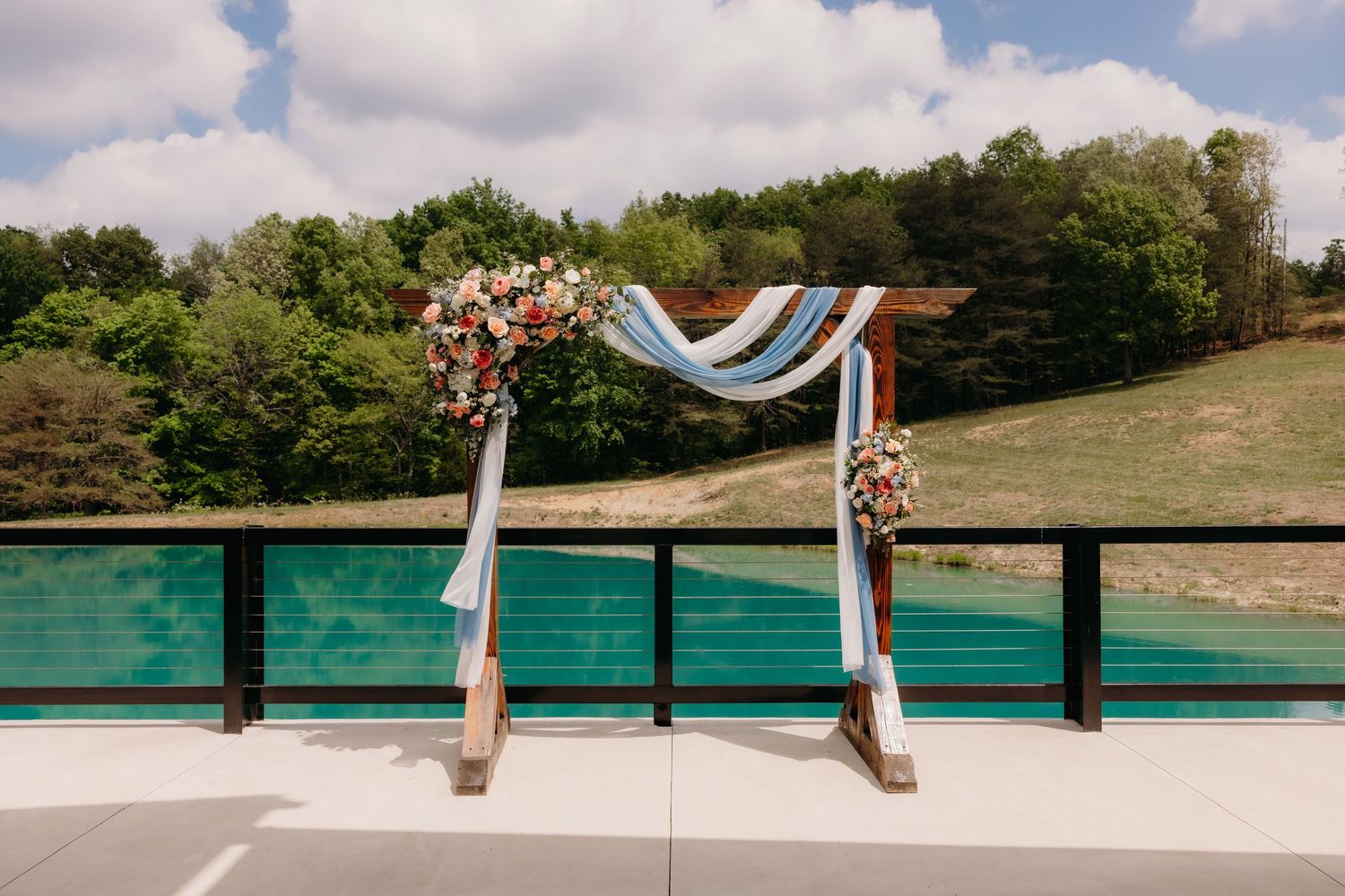 Wooden wedding arch adorned with pink flowers and blue draping on an outdoor deck overlooking a lush green hillside.