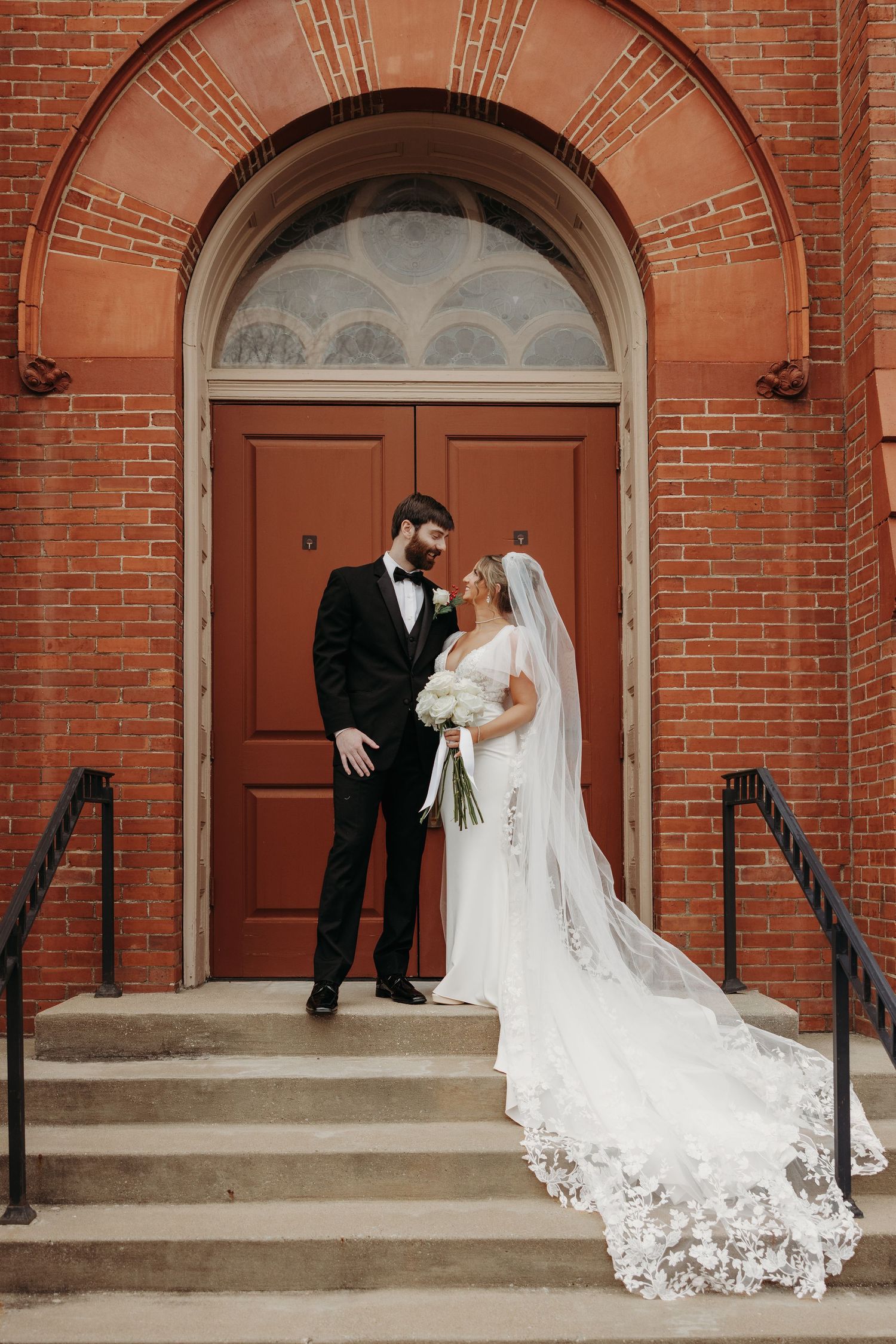 Bride and groom pose on church steps in front of red doors, bride wearing lace cathedral veil and white gown.