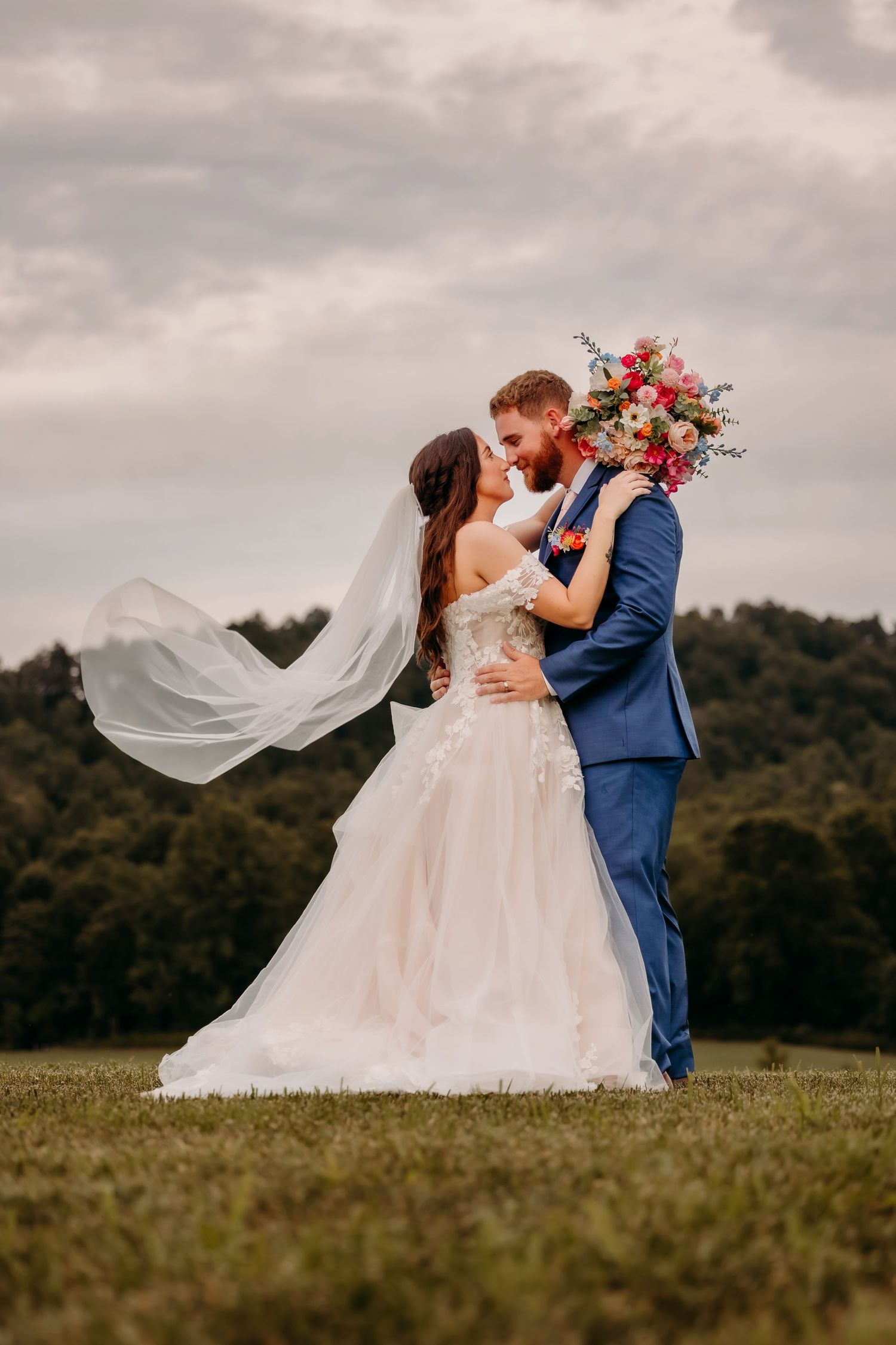 Bride and groom kiss outdoors with veil flowing in wind and colorful bridal bouquet.