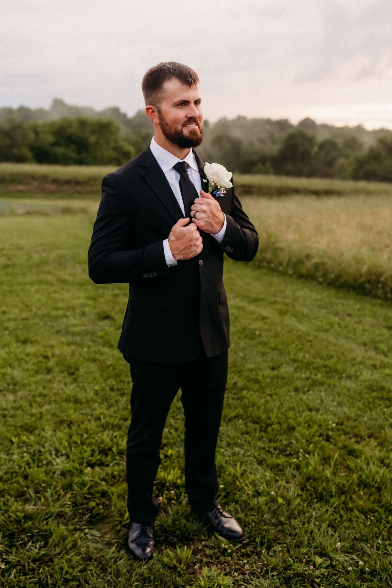 Bearded groom in black suit with white boutonniere adjusting jacket outdoors in green field.