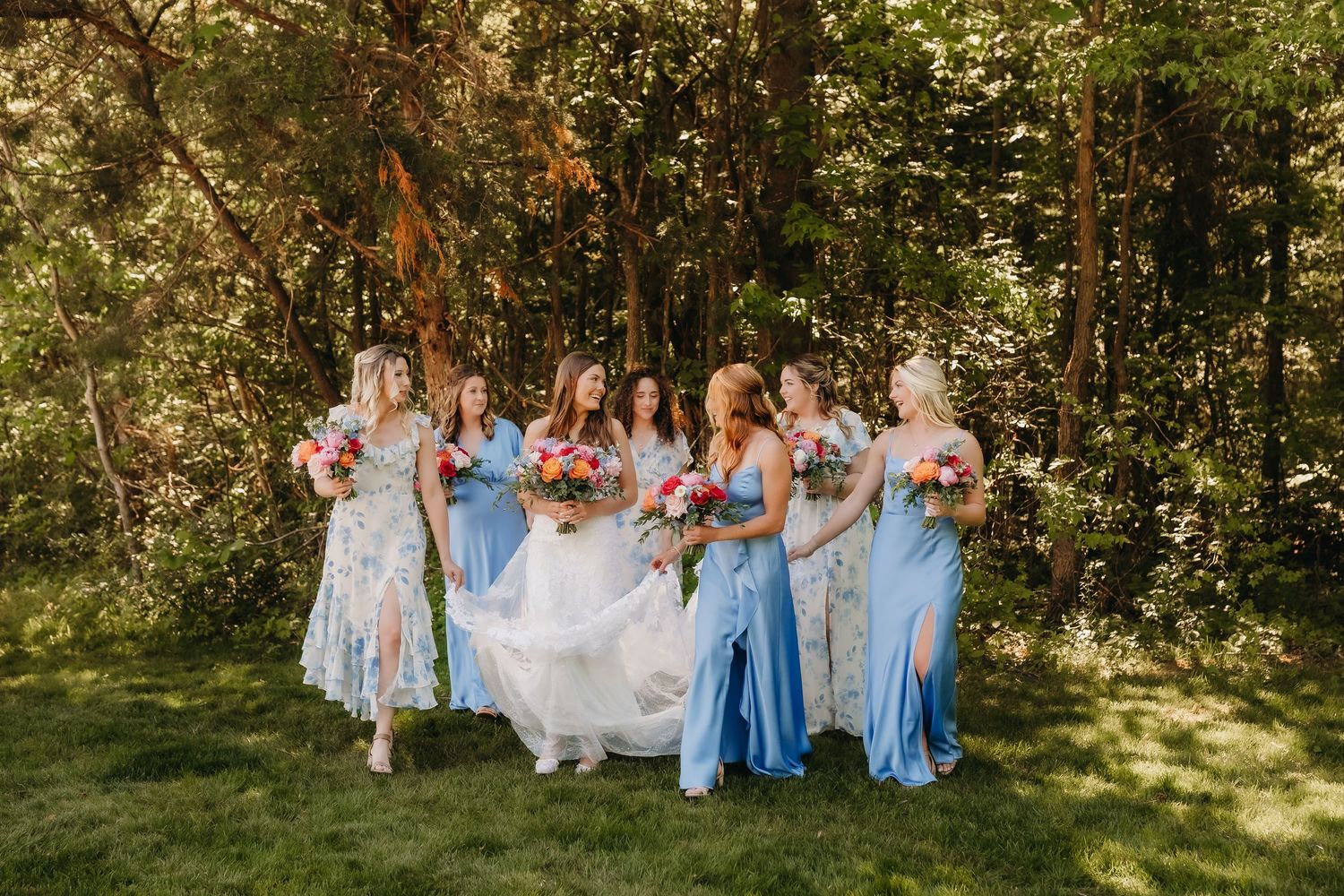 Bride in white gown with bridesmaids in blue dresses holding colorful bouquets outdoors in a lush green forest setting.