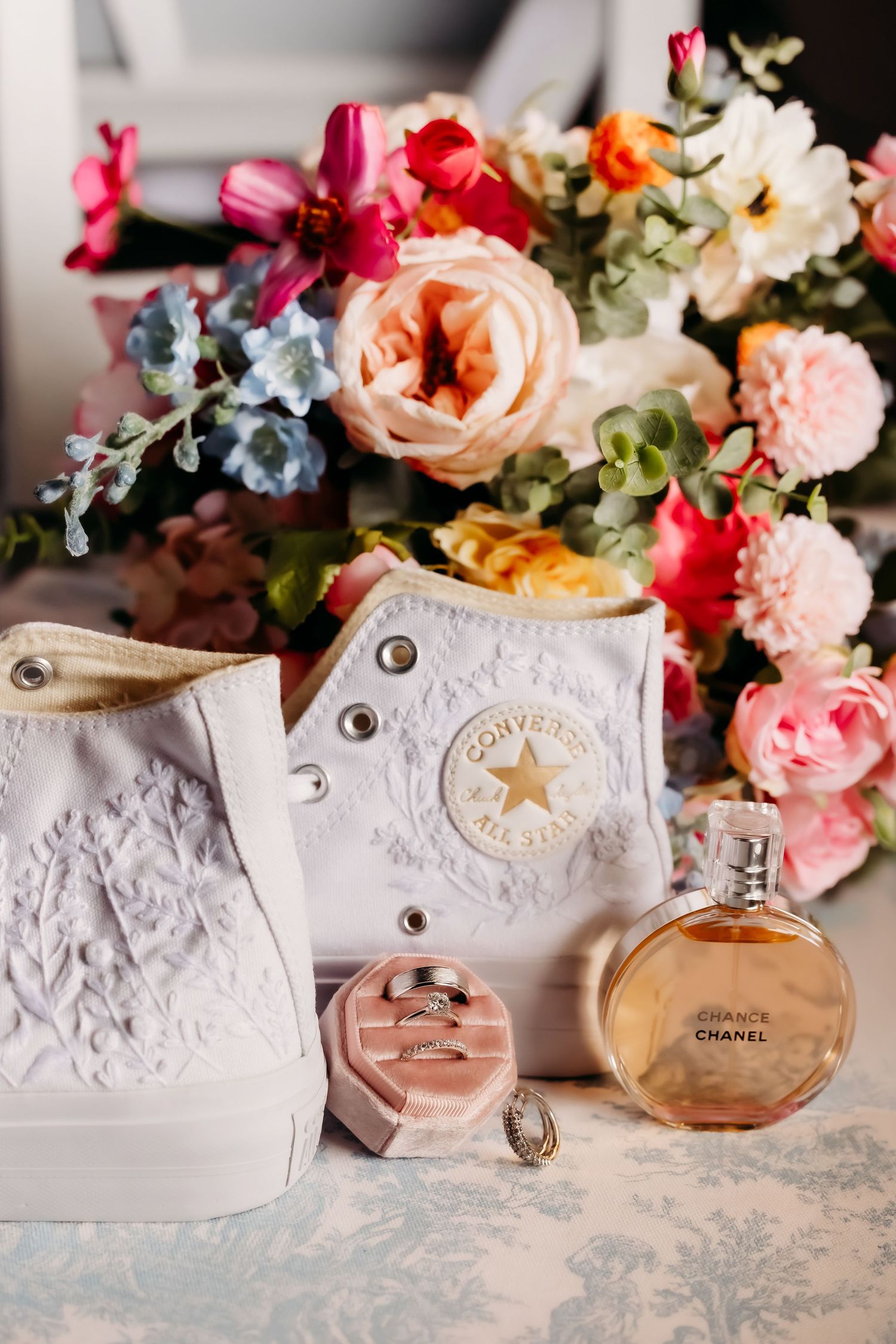 White Converse wedding shoes with lace detail, Chanel Chance perfume, pink ring box with rings, and colorful floral bouquet.