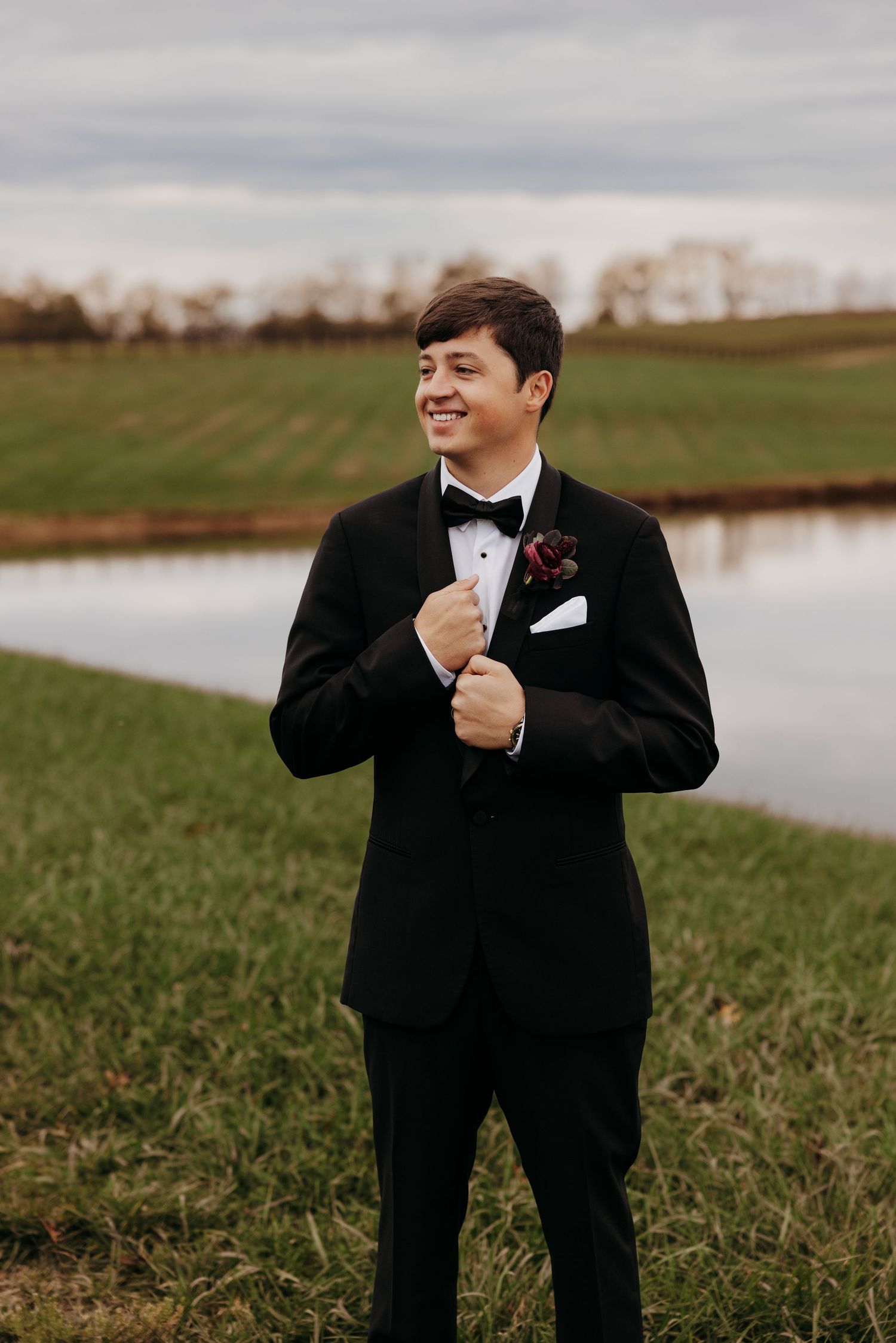 Smiling groom in black tuxedo with boutonniere standing near a pond in a green field.