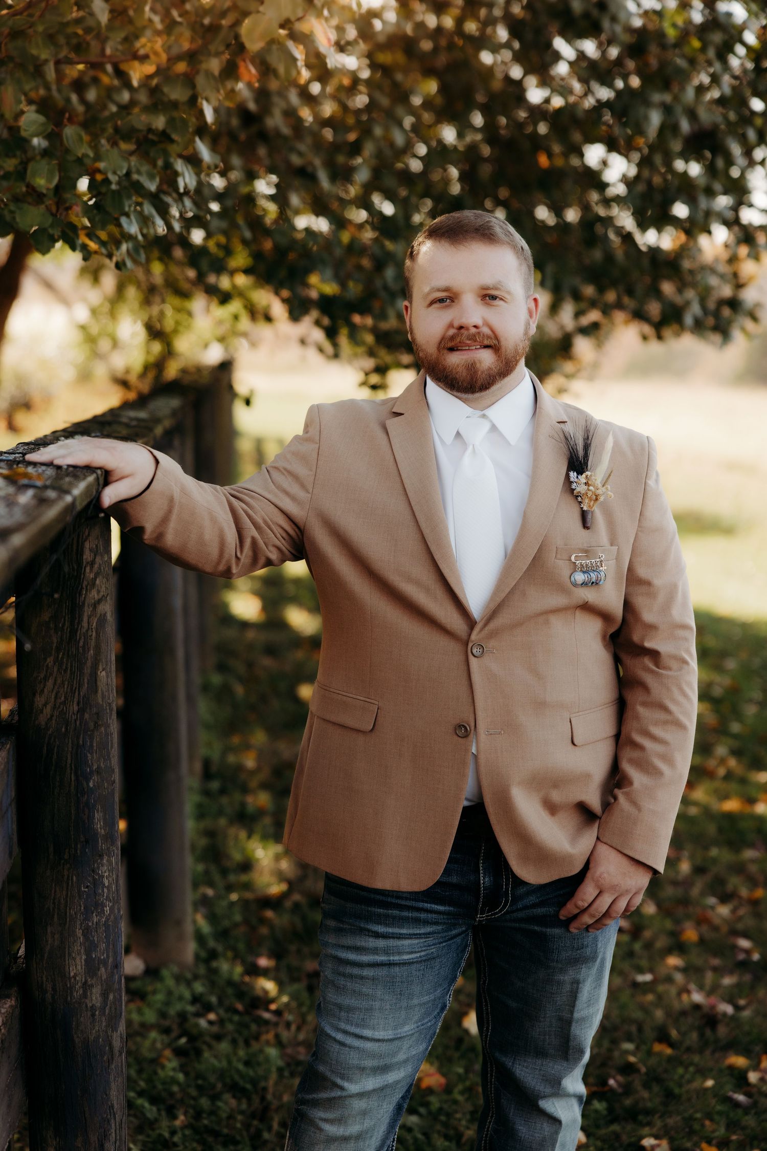 A bearded groom in a tan blazer, white shirt, and jeans leans on a wooden fence outdoors.