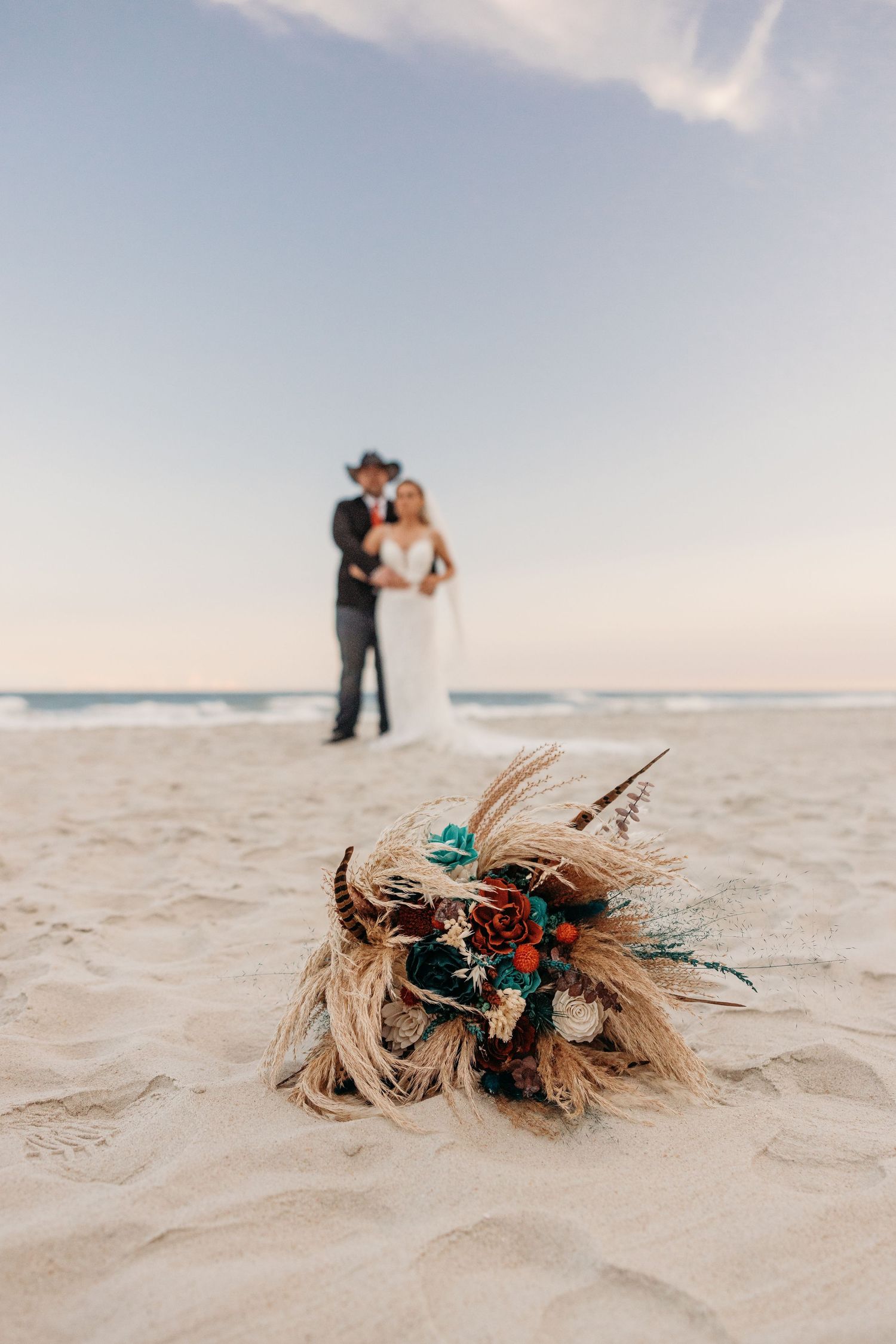 Wedding bouquet with turquoise accents lies in white sand with couple in background.