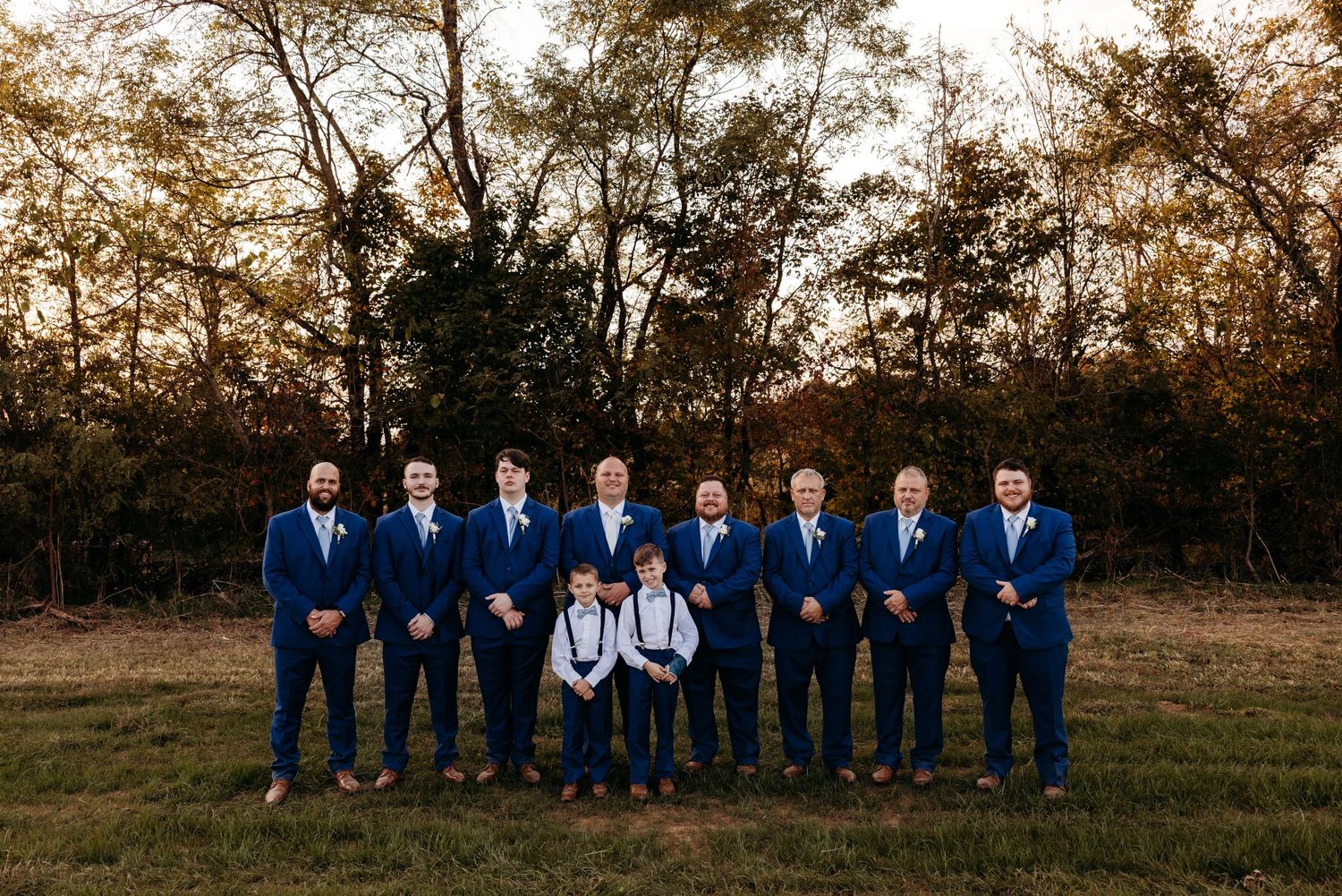 Groomsmen in navy blue suits with white boutonnieres pose outdoors with two young boys in matching attire.