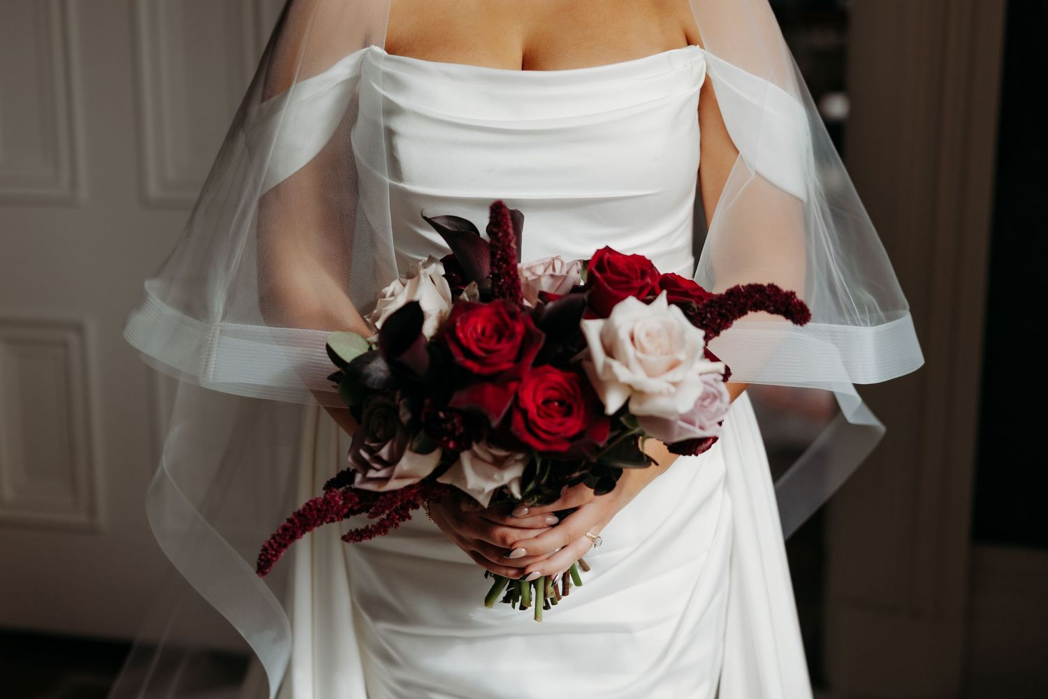 Bride in white strapless gown holding a bouquet of red and pink roses with dark foliage and a flowing veil.