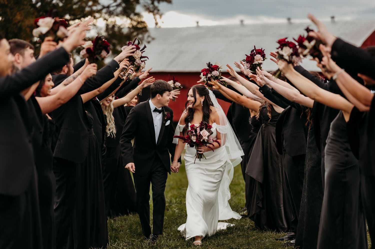 Bride and groom walk through arch of raised bouquets held by bridesmaids in black dresses at outdoor barn wedding.