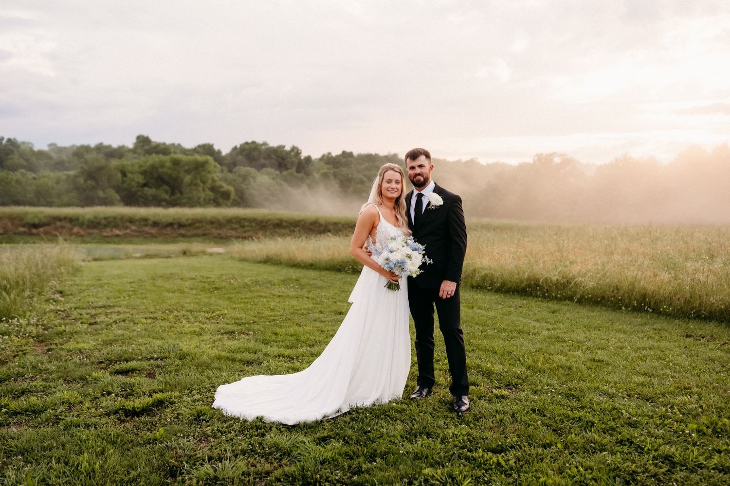 Bride and groom posing together in a lush green field at golden hour sunset on their wedding day.