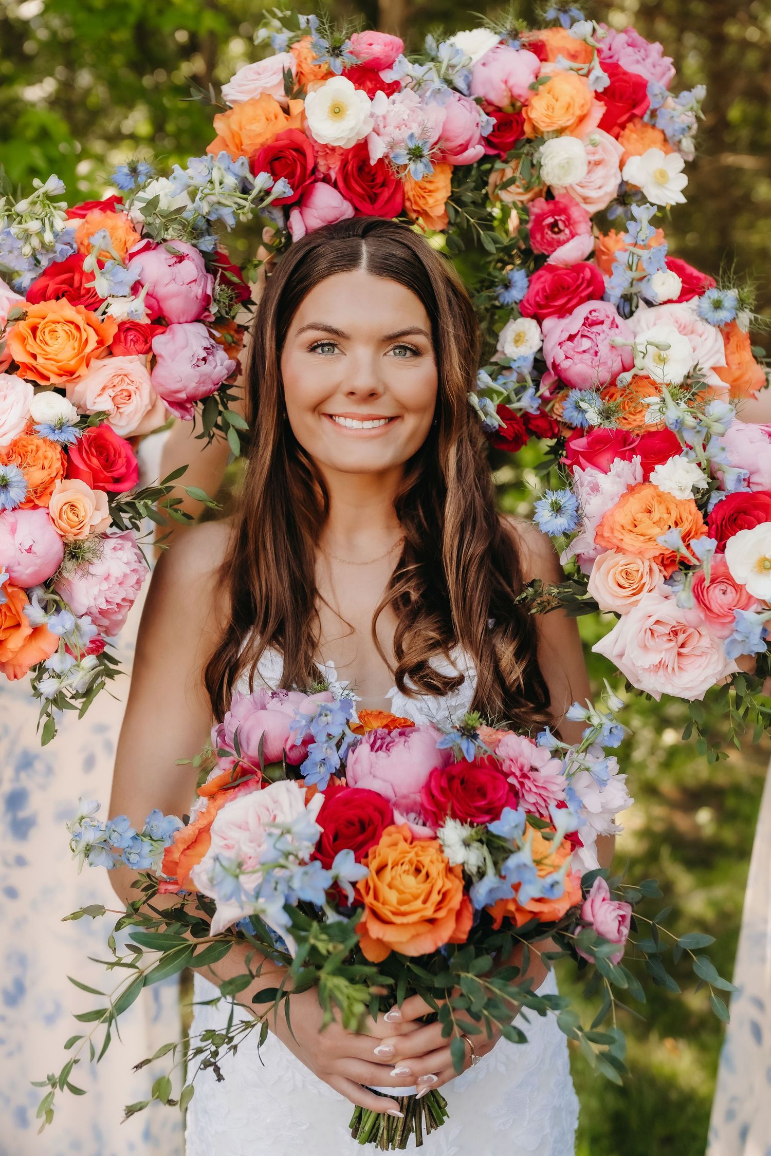 Smiling bride holding vibrant bouquet framed by large colorful floral arch with peonies, roses, and blue blooms.
