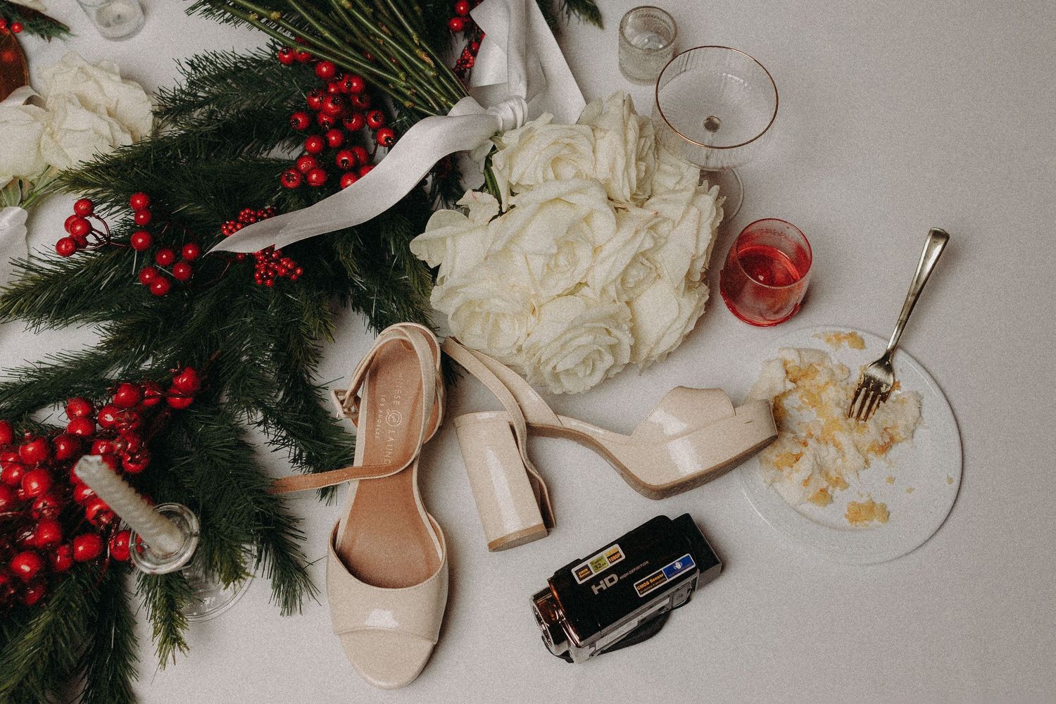 Flat lay of bridal accessories including nude heels, white floral bouquet, holly berries, cake slice, and vintage camera on white surface.