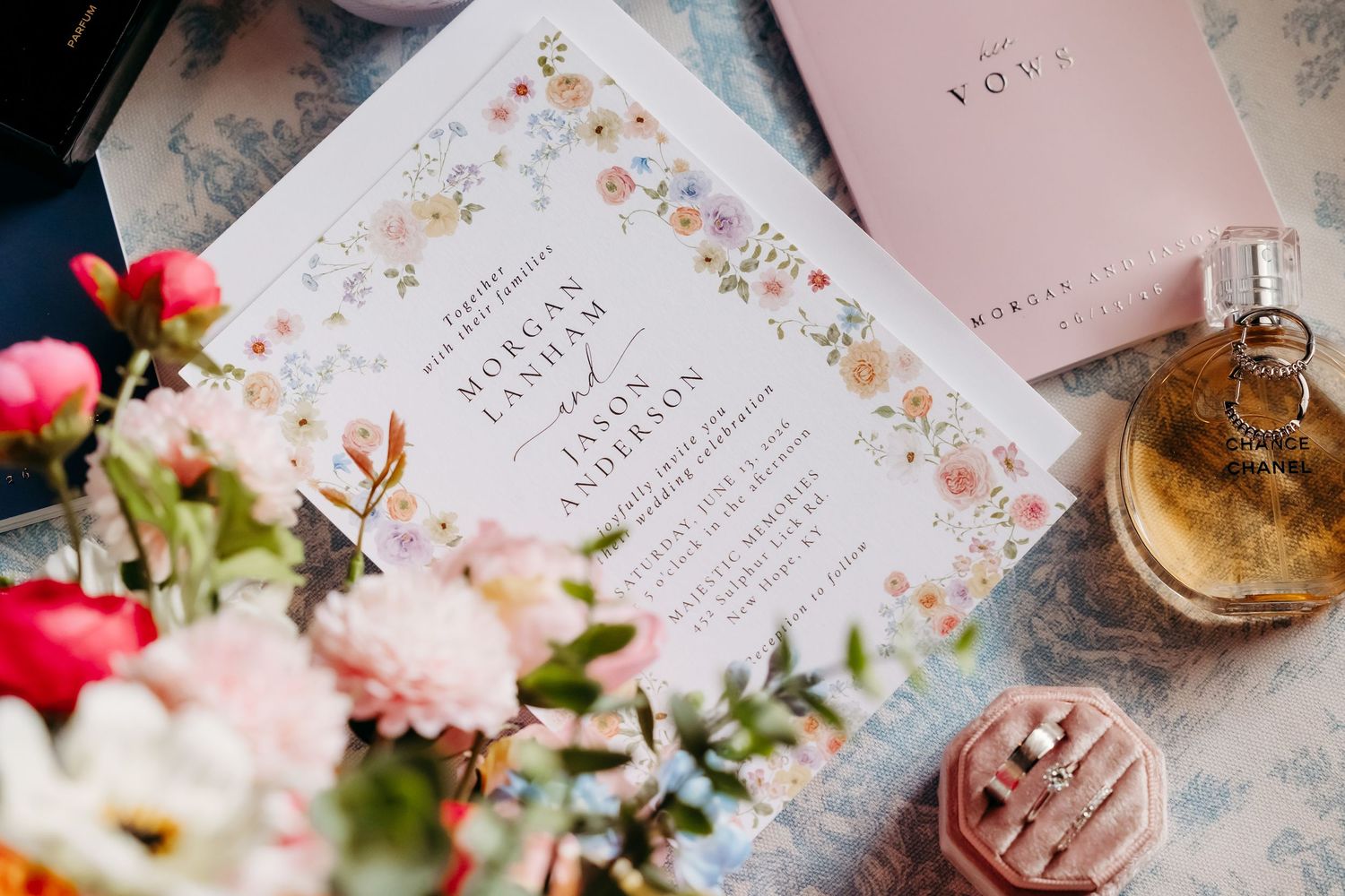 Floral wedding invitation with colorful wildflower border beside a bouquet, perfume bottle, and ring box on a blue surface.