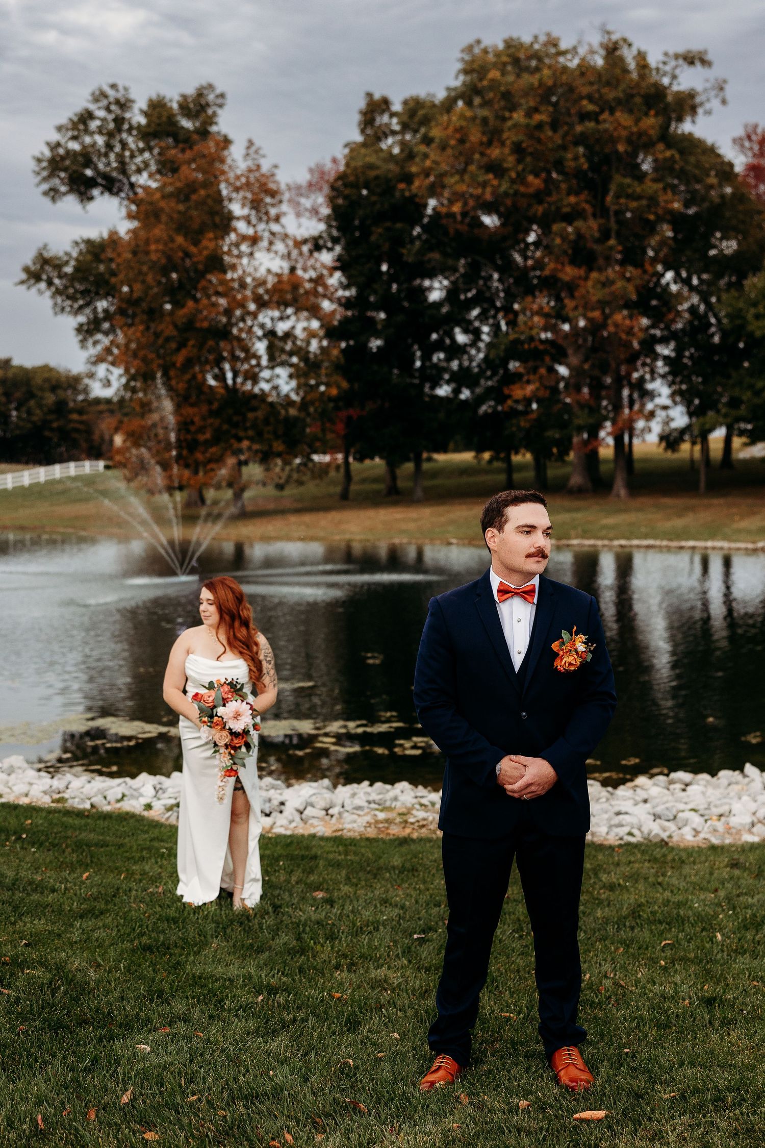 Bride and groom posing by a pond with autumn trees during outdoor wedding photo session.