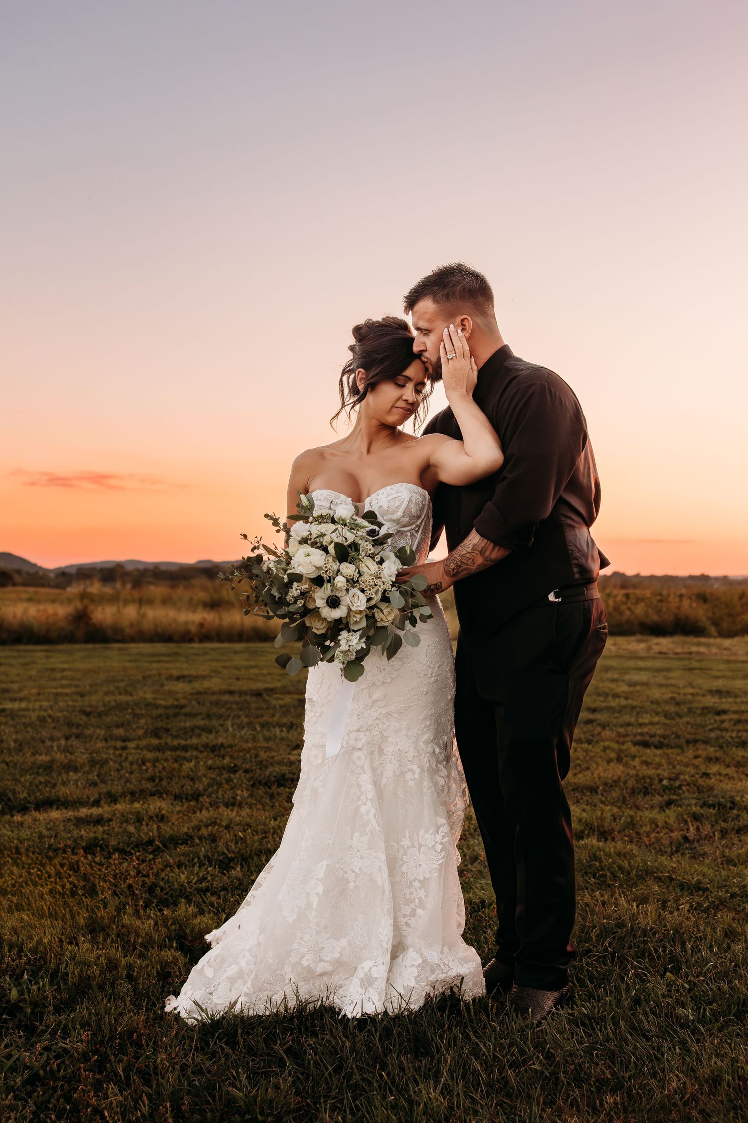 Bride and groom embrace at sunset in a field, bride in lace gown holding white floral bouquet.