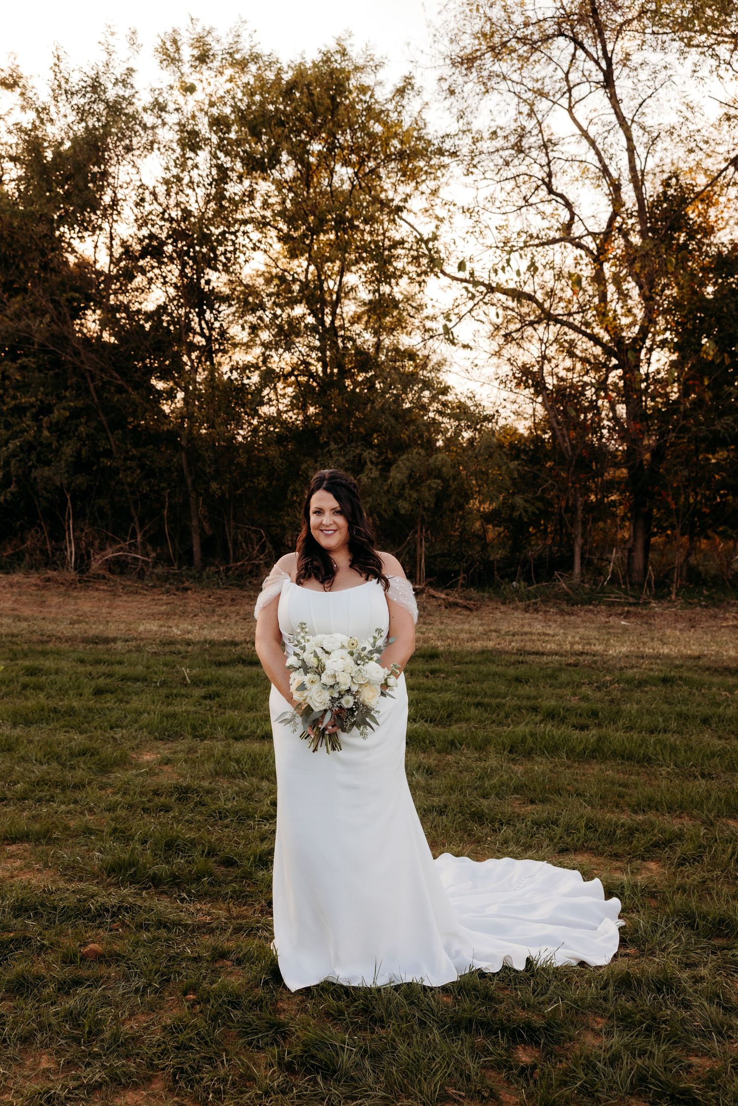 Bride in fitted white gown holding bouquet standing in golden hour sunlit field.