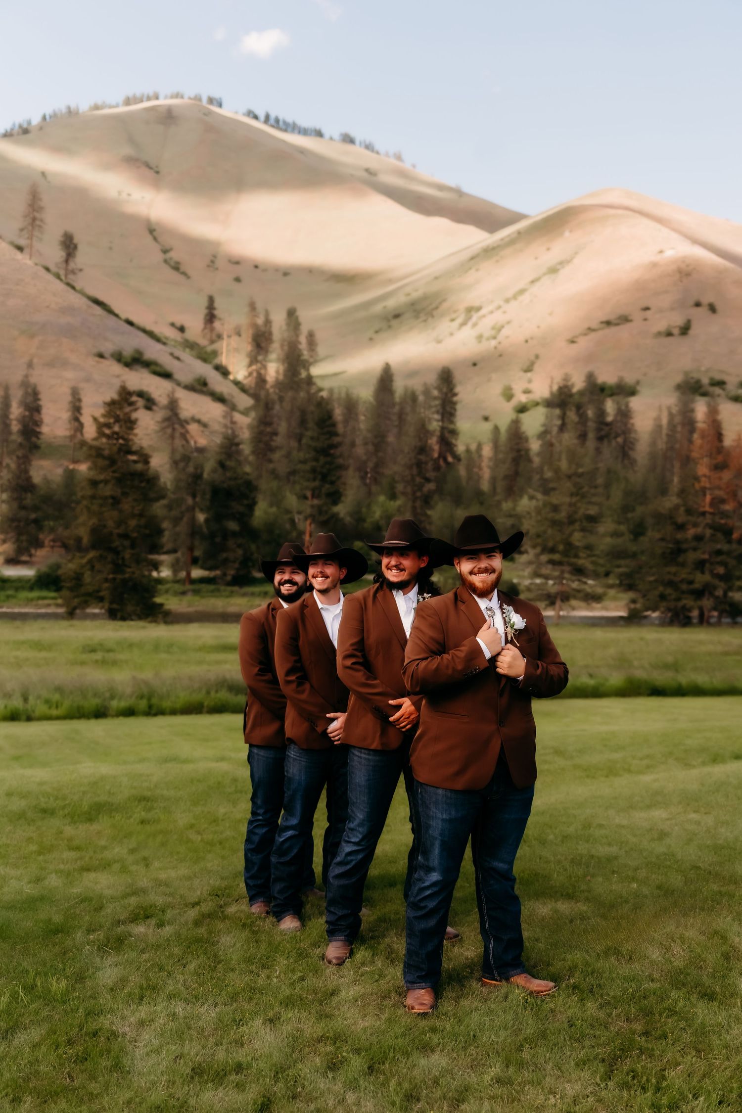 Groom and groomsmen in brown blazers and black cowboy hats pose on green grass with mountain views.