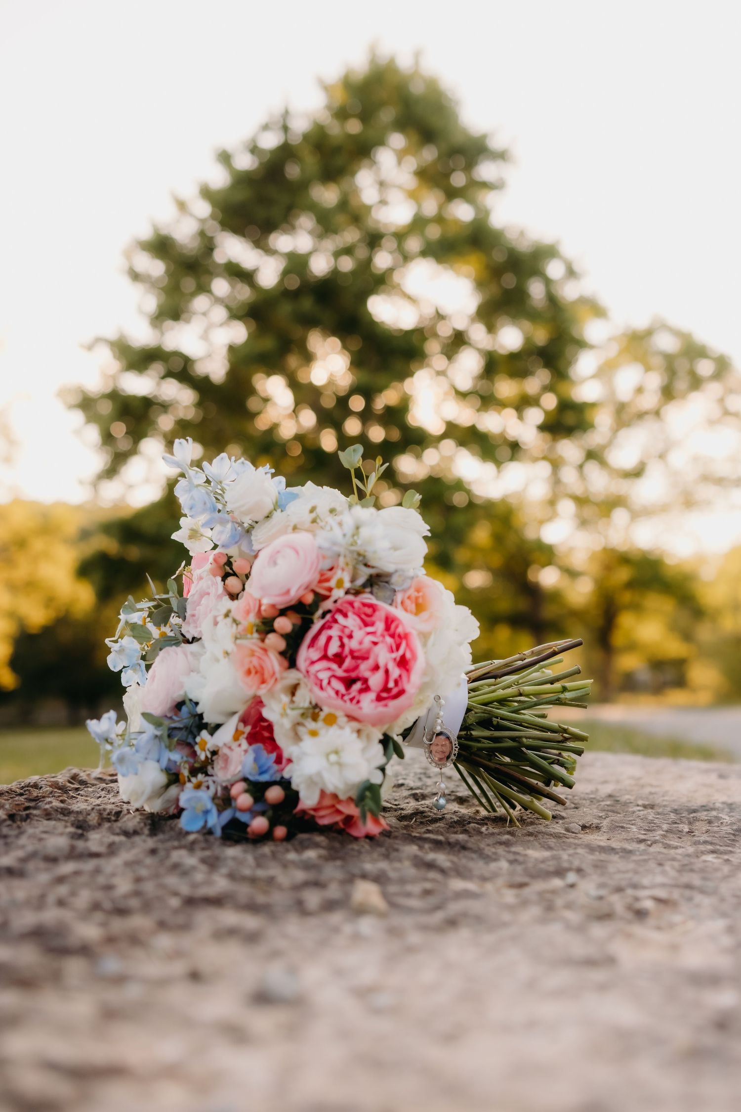 Bridal bouquet with pink peonies, white roses, and blue flowers resting on stone at sunset.