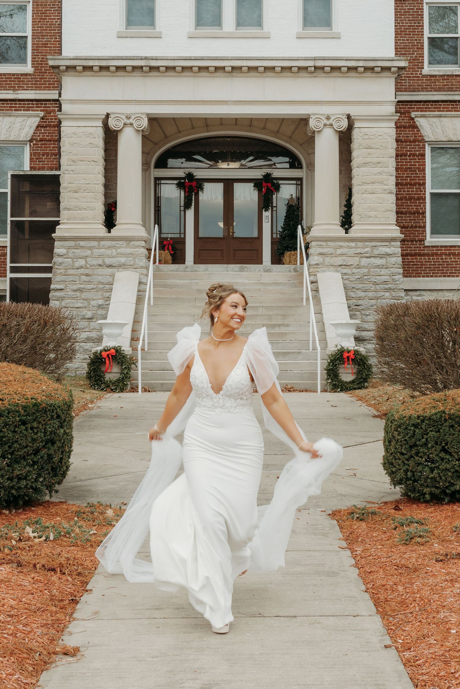 Radiant bride in white V-neck wedding gown with sheer sleeves walks outside decorated brick building in winter.