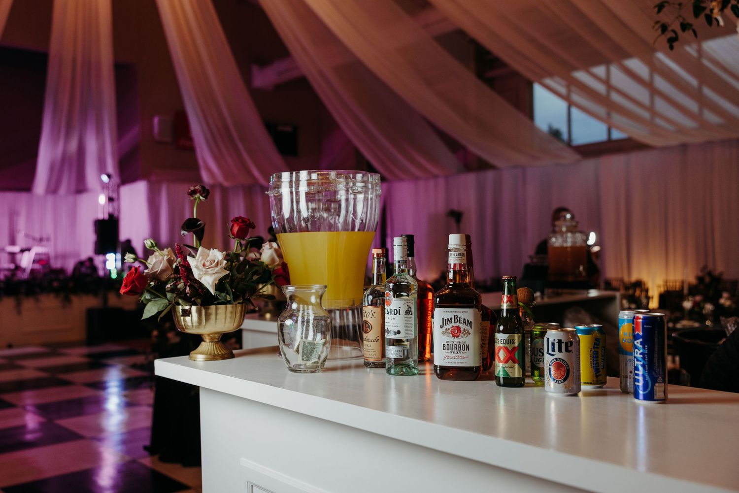 Wedding bar setup with orange juice pitcher, assorted liquor bottles, and floral decor in a draped venue.