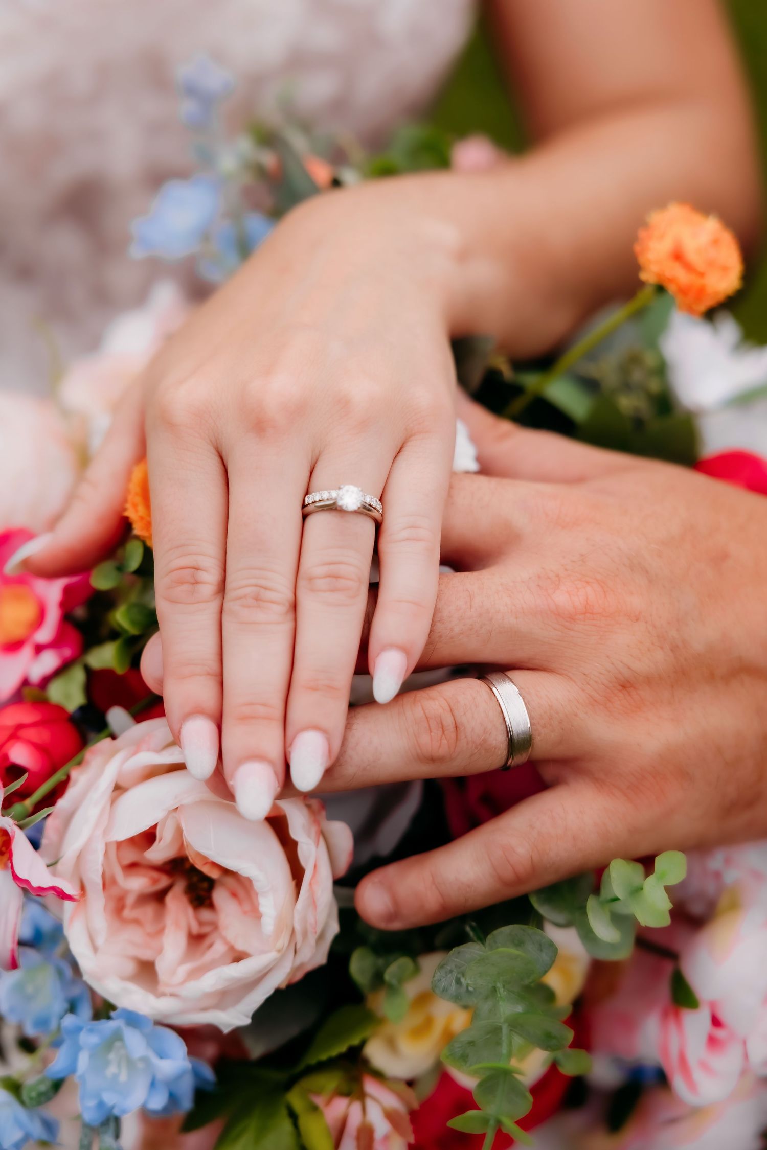 Bride and groom display wedding rings over a vibrant colorful bridal bouquet of flowers.
