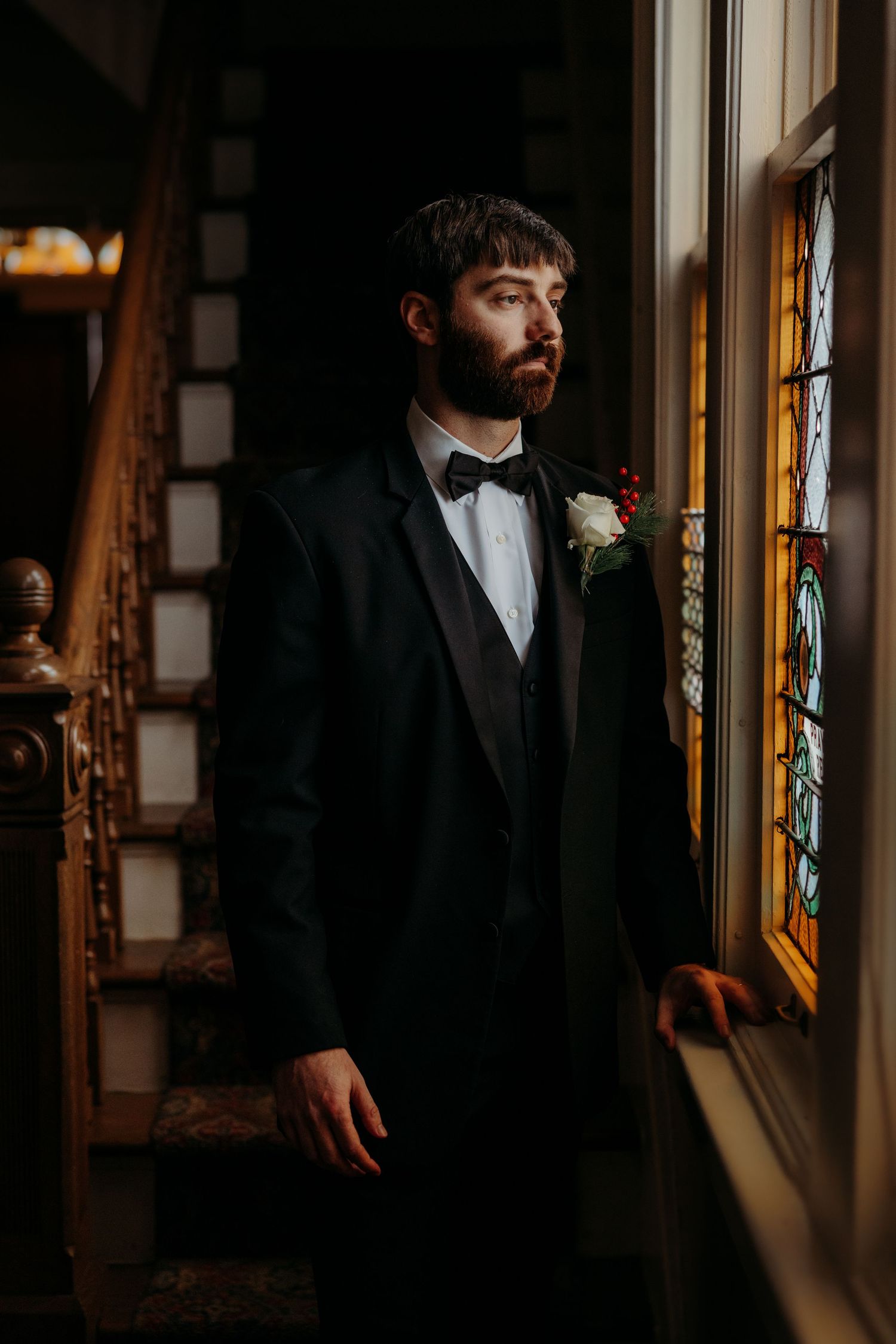 A groom in a classic black tuxedo poses near a stained glass window.