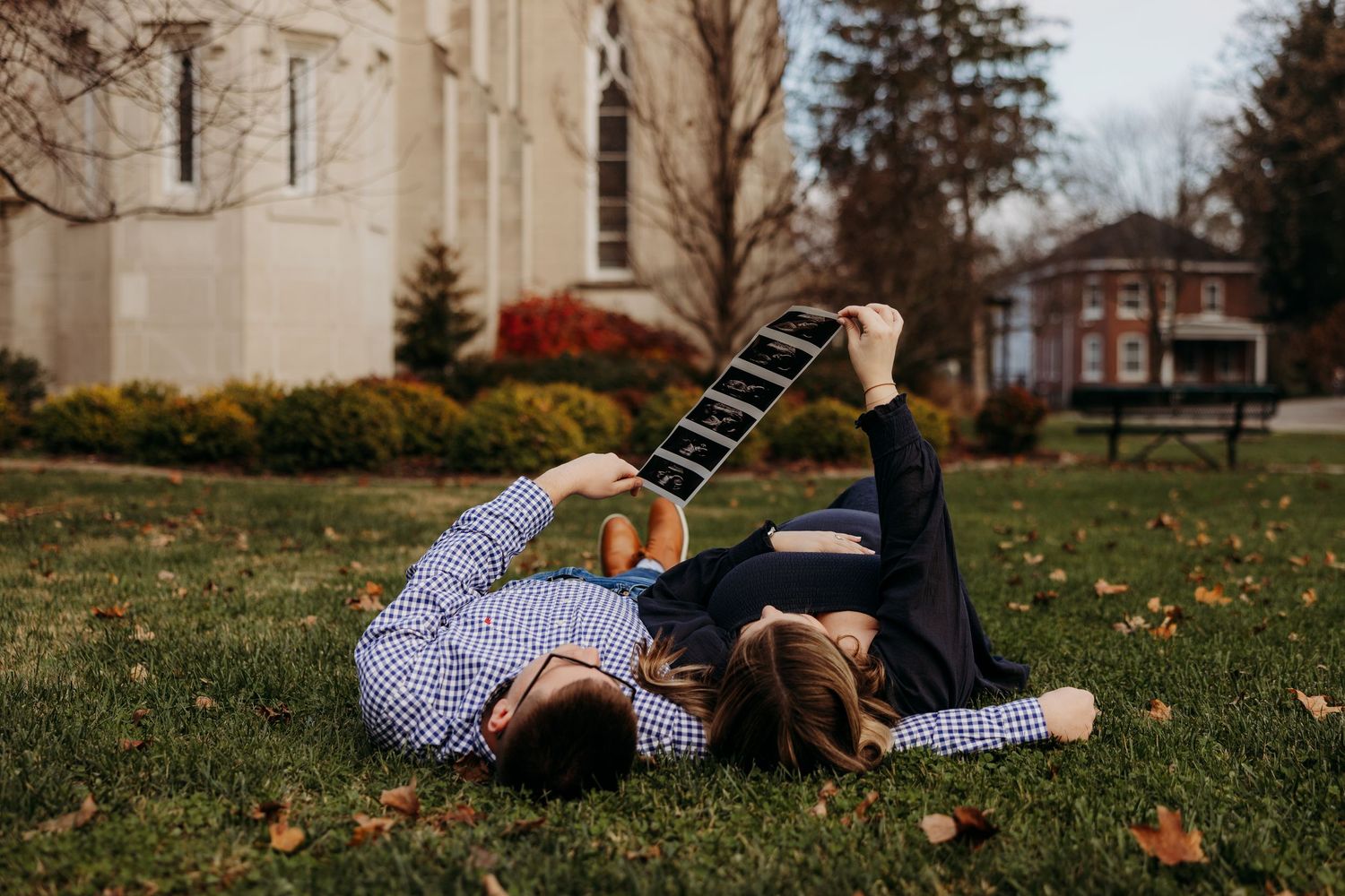 Couple lying on grass playing guitar near historic brick building with autumn leaves in background.