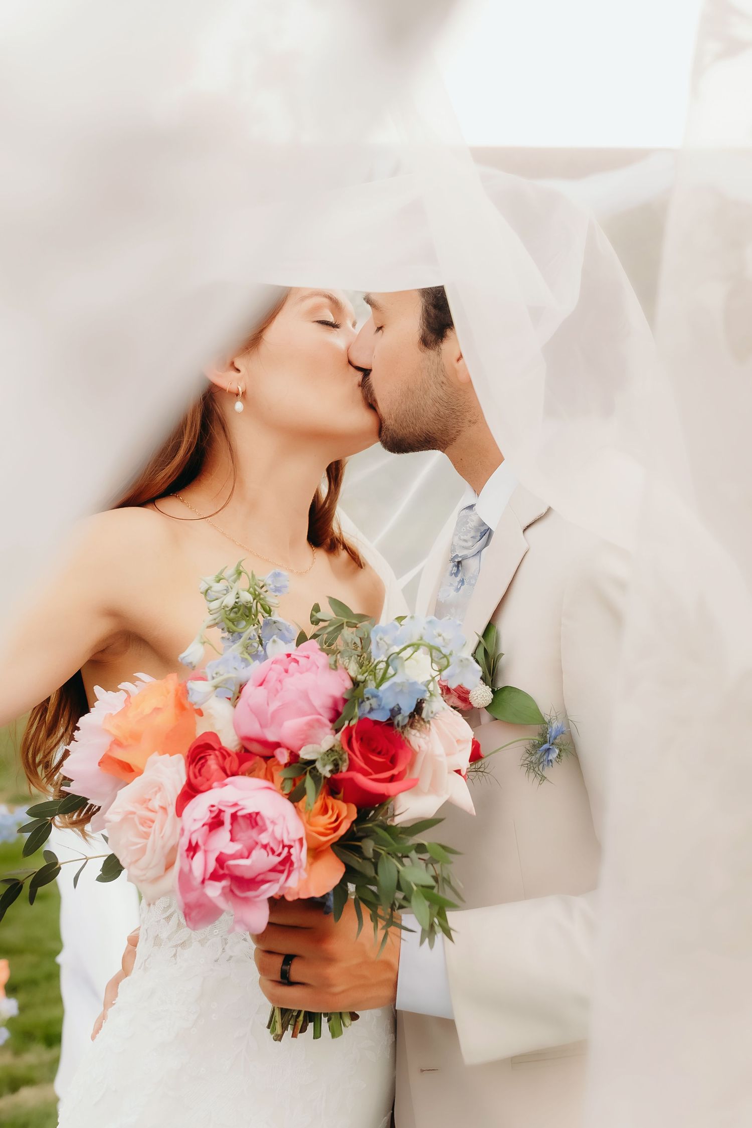 Bride and groom kiss under a veil as bride holds a vibrant pink and coral peony wedding bouquet.