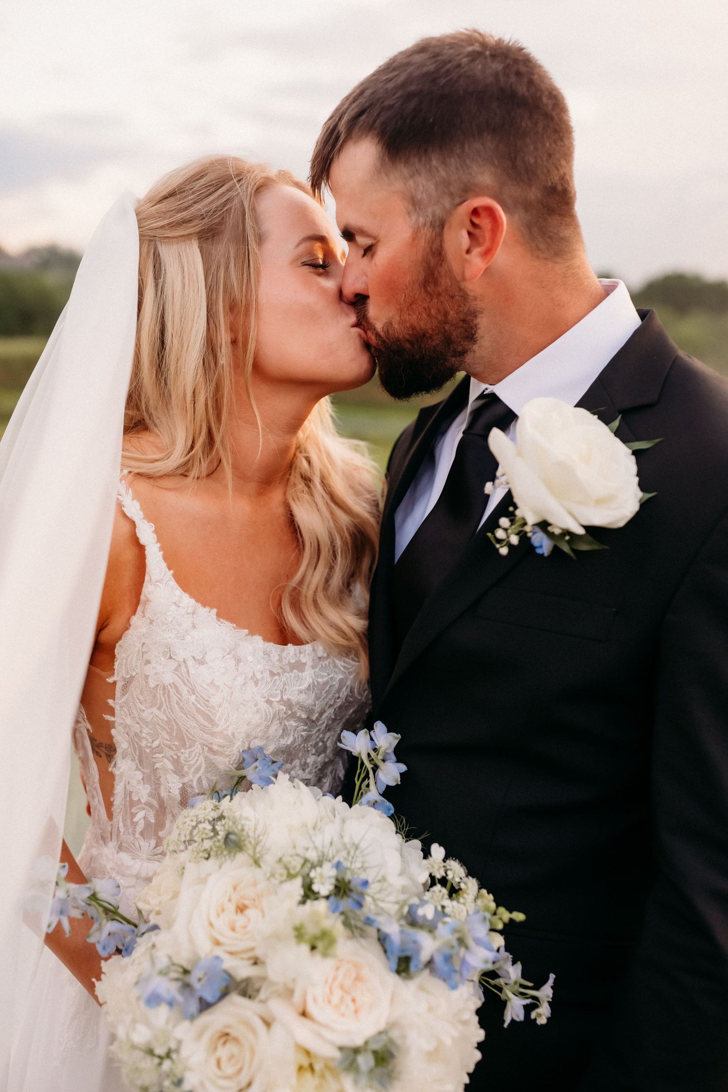 Bride and groom sharing a romantic kiss, bride holding white and blue floral bouquet in lace wedding dress.