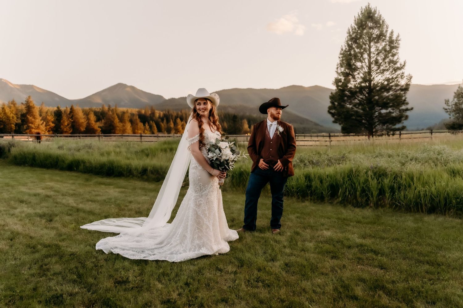 Bride in white gown and cowboy hat stands with groom in western attire on green meadow with mountain backdrop.