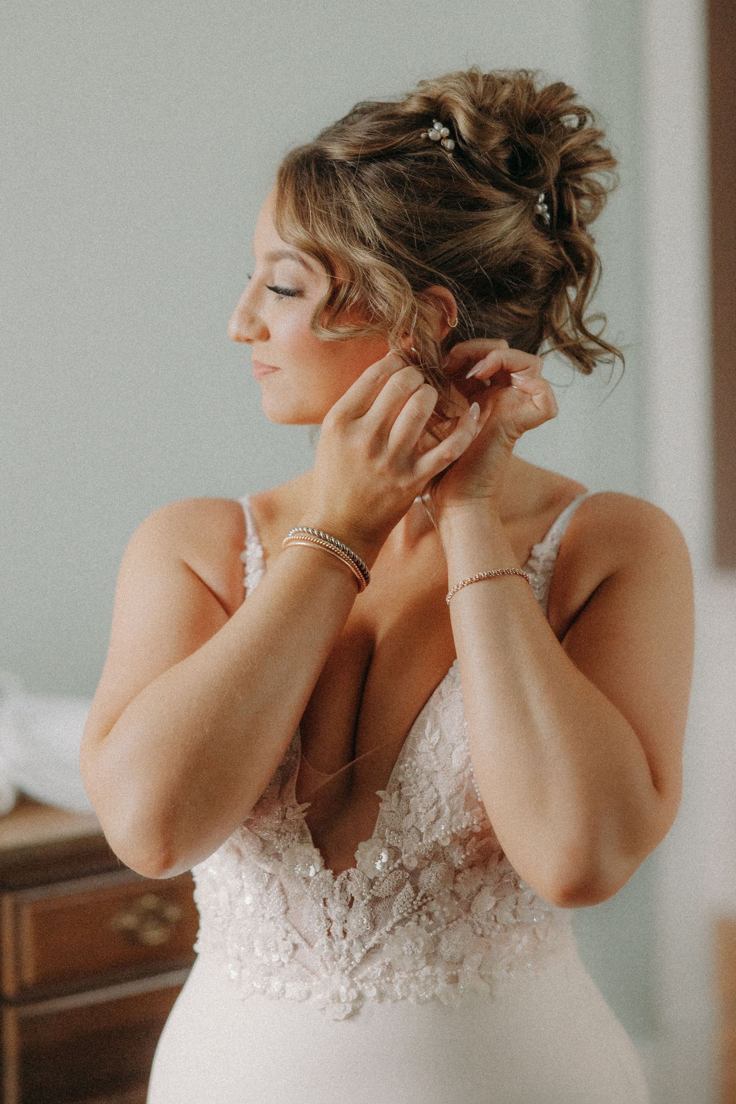 Bride in lace wedding dress putting on earrings, hair styled in elegant updo with pearl bracelet.