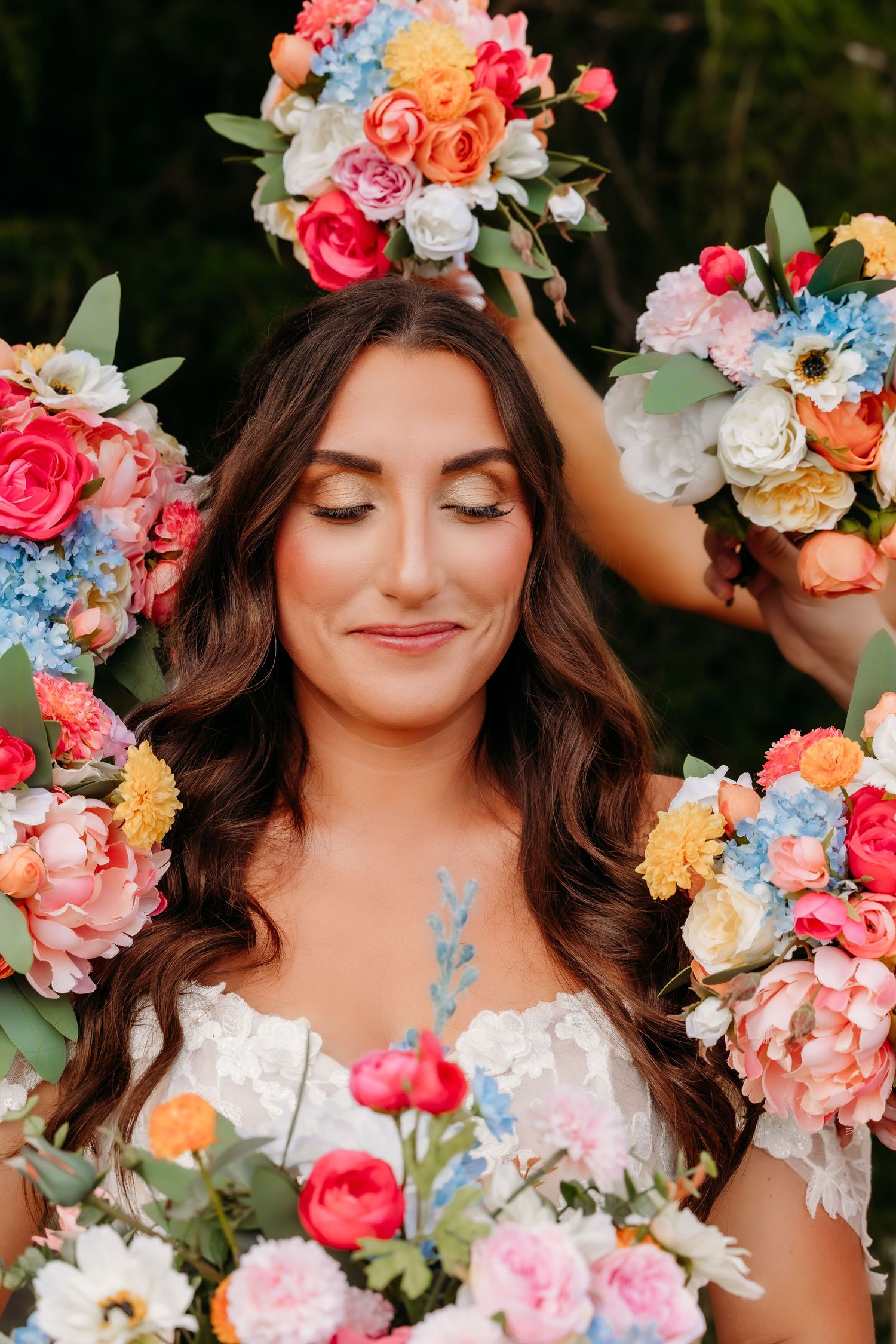 Bride surrounded by colorful floral bouquets with eyes closed, wearing a white lace dress.